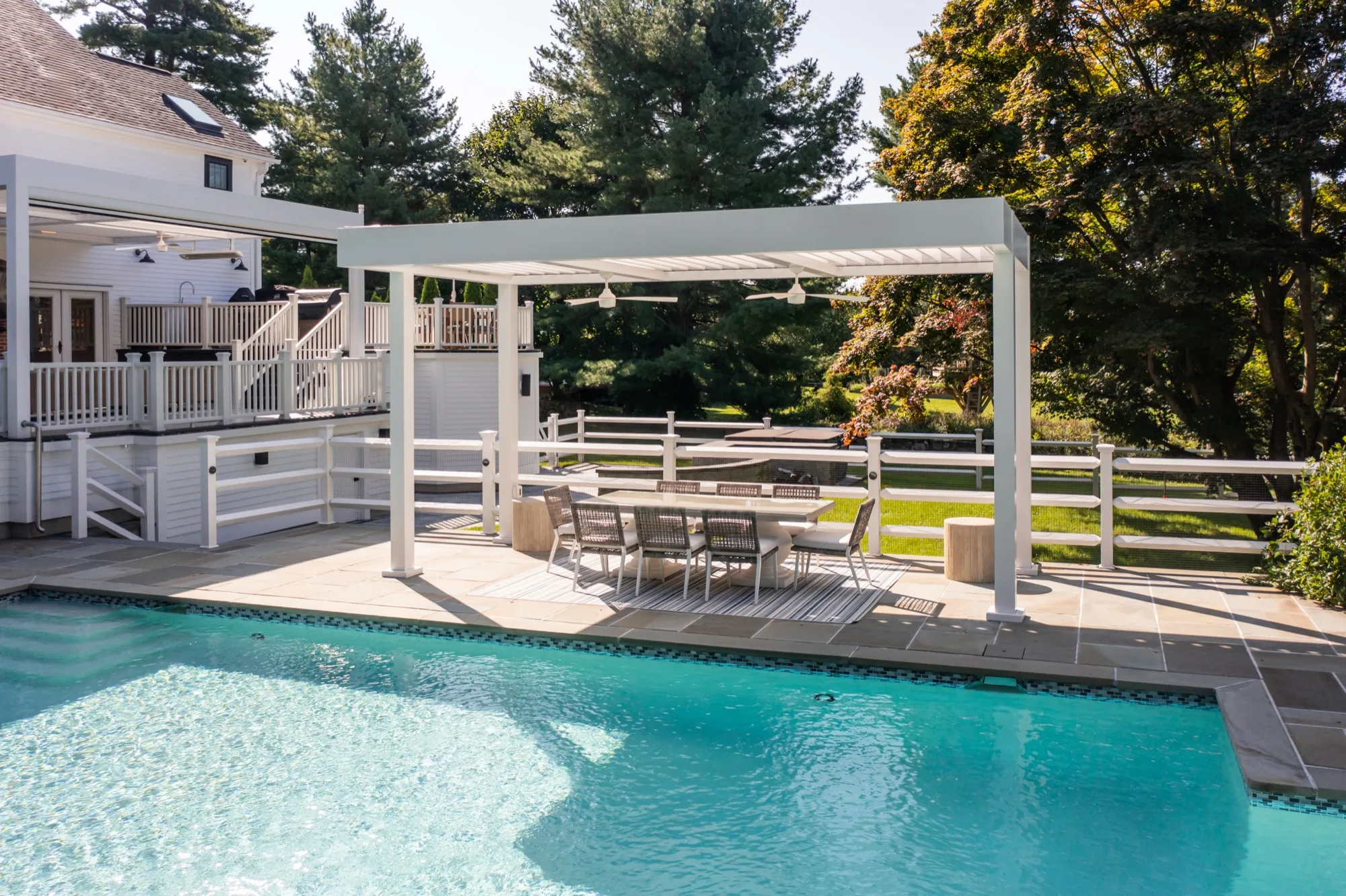 Daytime aerial photo of a pool with a louvered pergola shading a dining set on the adjoining patio, autumn foliage visible