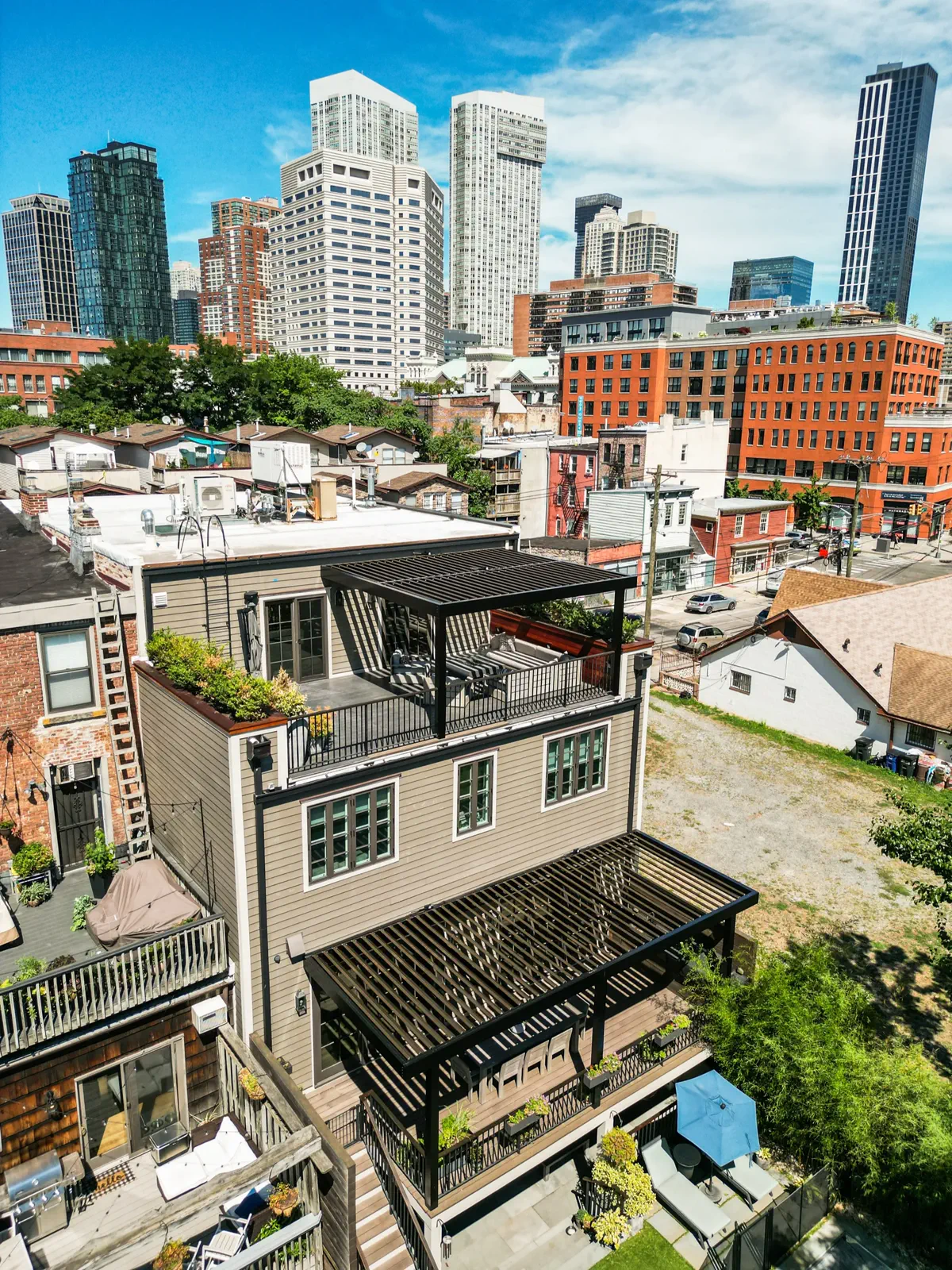 Daytime aerial photo of a rooftop deck with a black louvered pergola atop a city rowhouse, Manhattan skyline in background