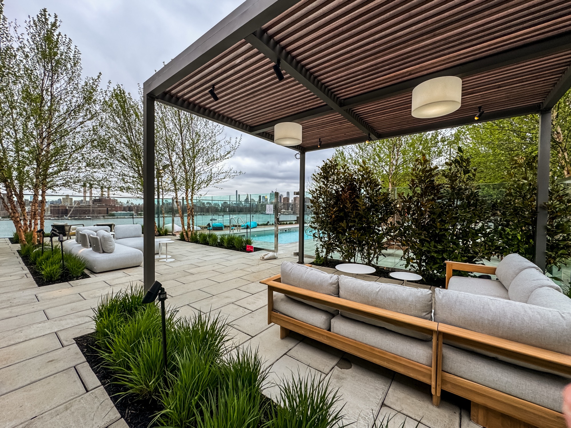Overcast daytime photo of a rooftop lounge with a wood-slat pergola, white sofas, a pool, and a city skyline across the water