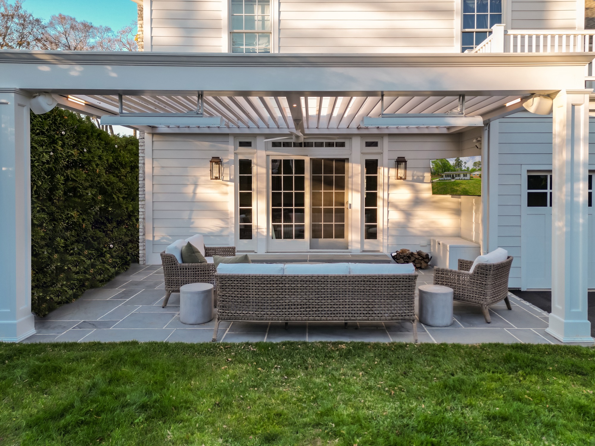 Shaded backyard patio with wicker furniture under a white louvered pergola attached to a house