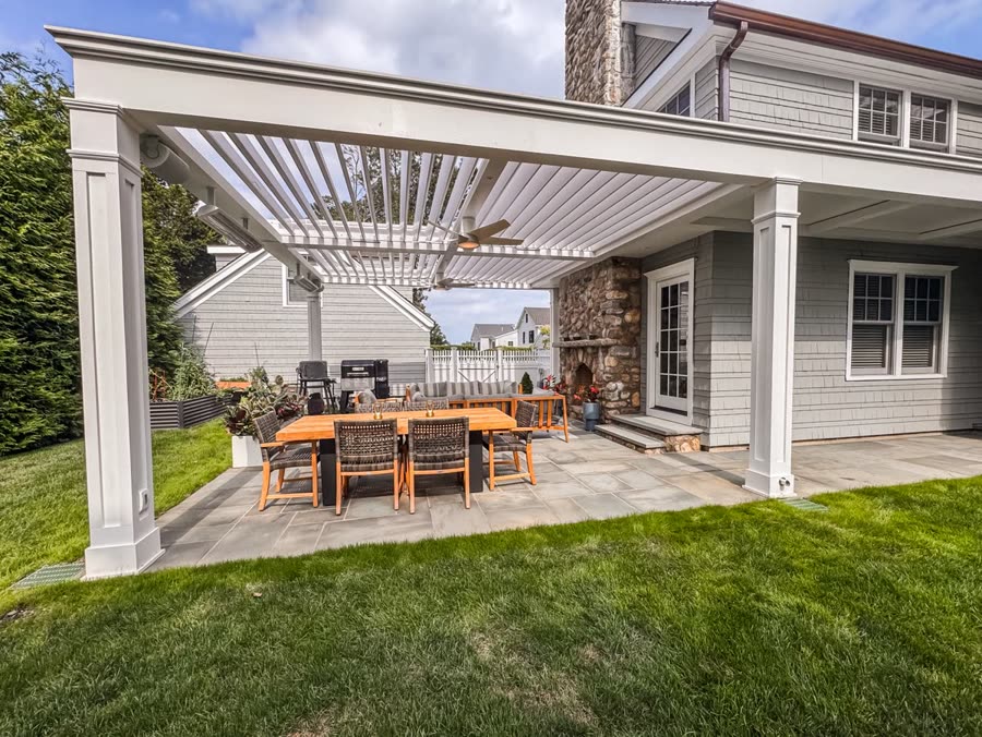 A white louvered pergola over a stone patio with a wood dining table and chairs