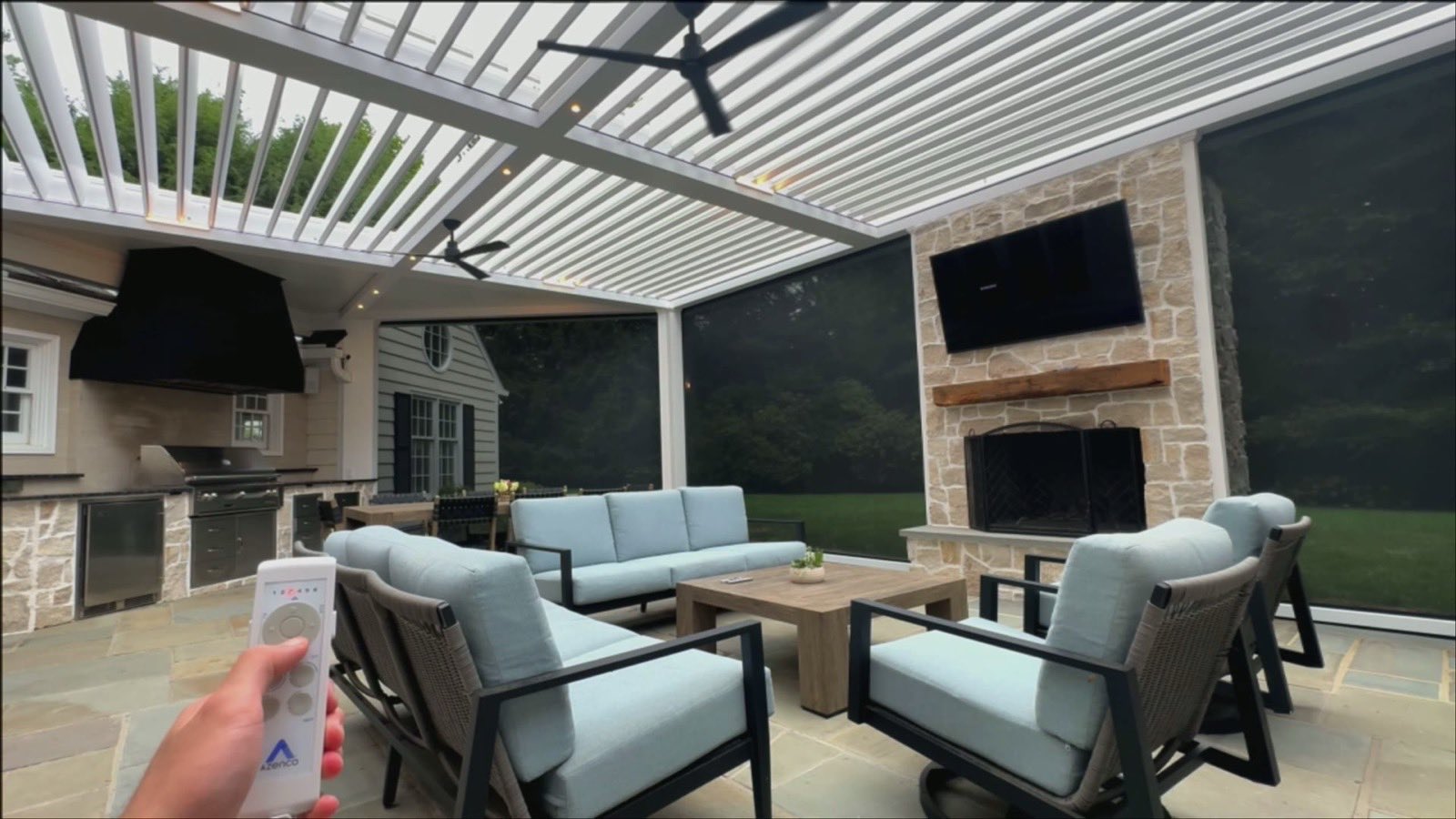 Photo of a covered patio with dark motorized screens down, stone fireplace, TV, and a hand holding an Azenco remote, daytime.