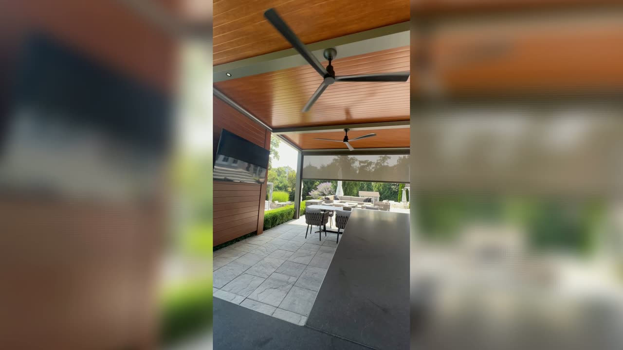 Photo of a wood-toned pergola ceiling with a fan and partially lowered motorized screen over a stone patio, daytime