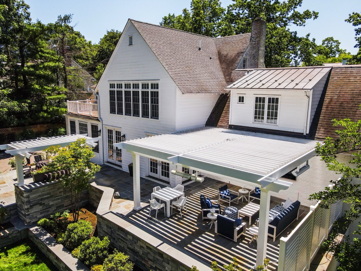 Aerial photo of a white louvered pergola attached to a large shingle-style house over a stone patio with navy furniture