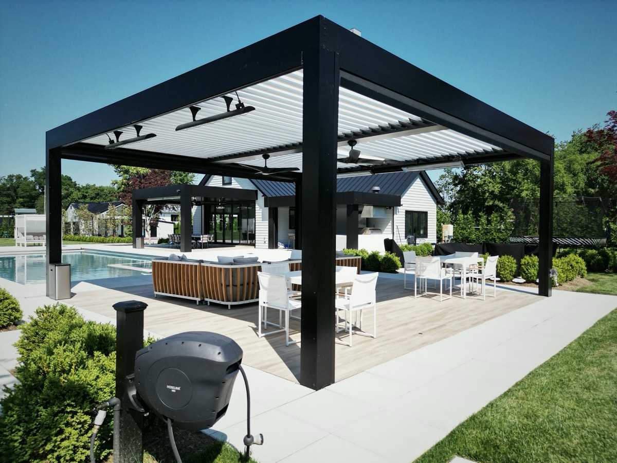A black freestanding louvered pergola over a pool deck patio with white and wicker furniture