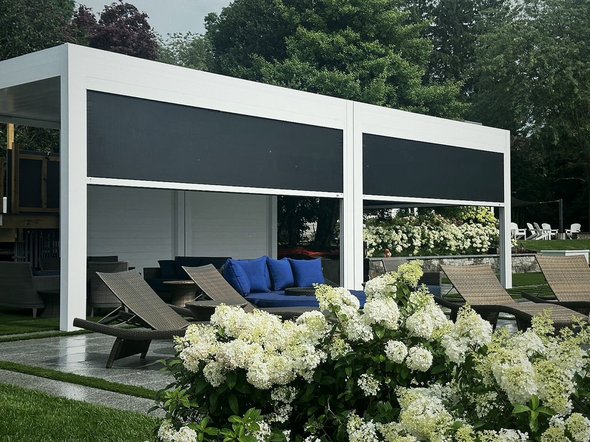 White pergola with closed black retractable screens beside wicker loungers and white hydrangeas