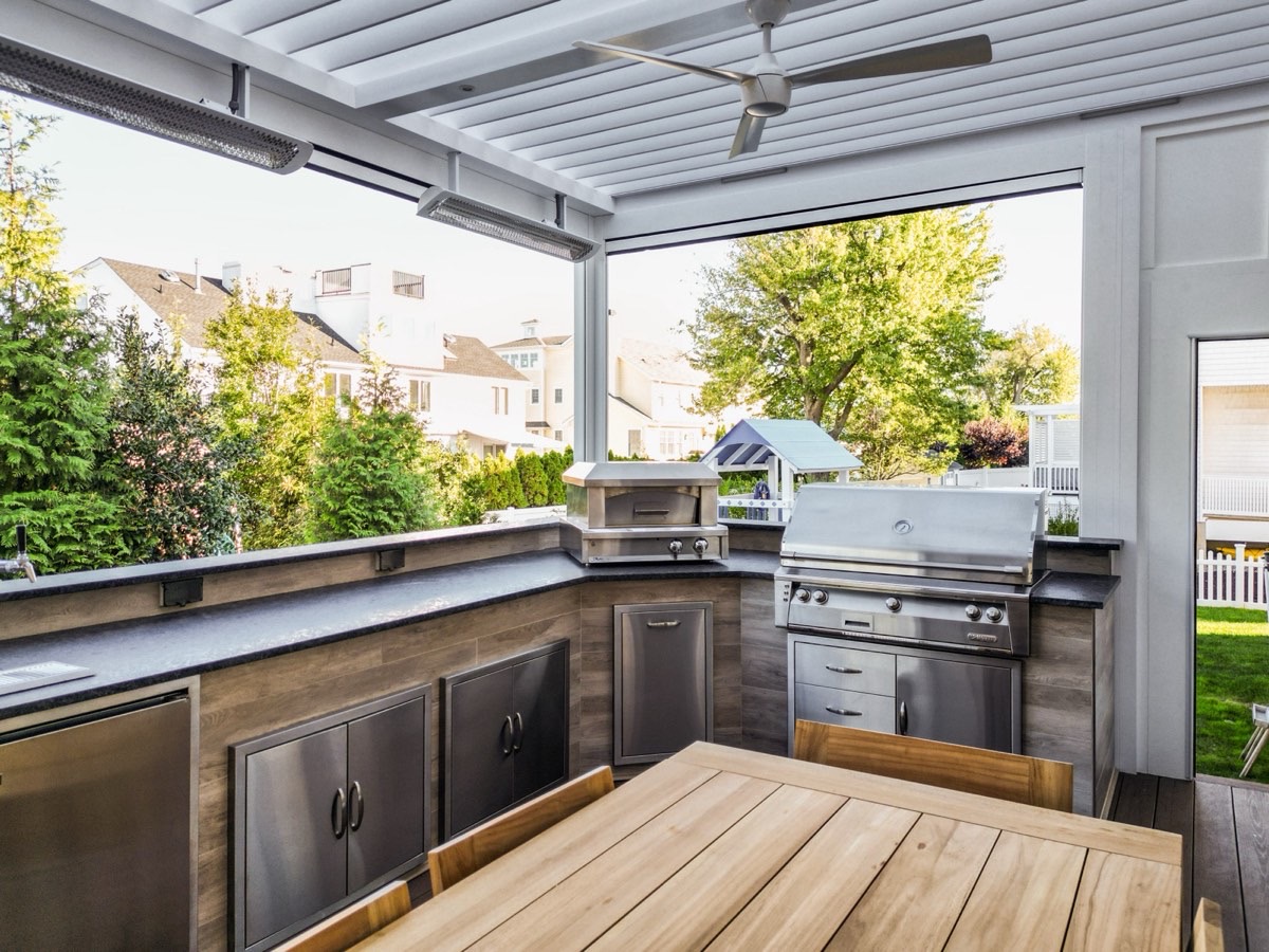 Pergola-covered outdoor kitchen with grill, pizza oven, and wood cabinetry, neighboring houses visible