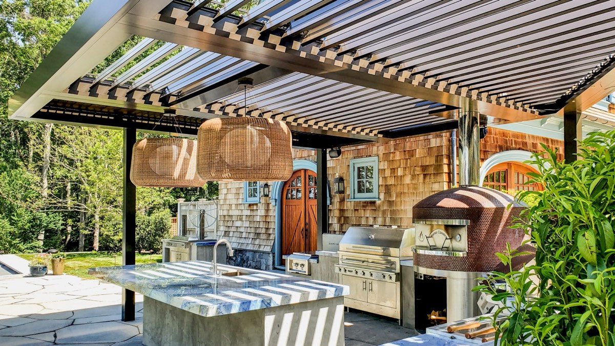 Photo of an outdoor kitchen with grill, sink island, and pizza oven beneath a dark slatted pergola roof, daytime, summer