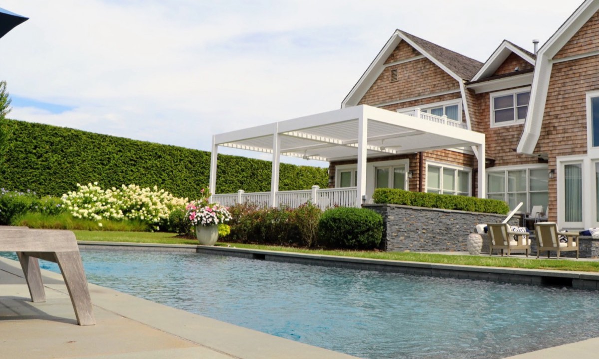 A white pergola attached to a shingle-style house beside a swimming pool in Southampton, NY