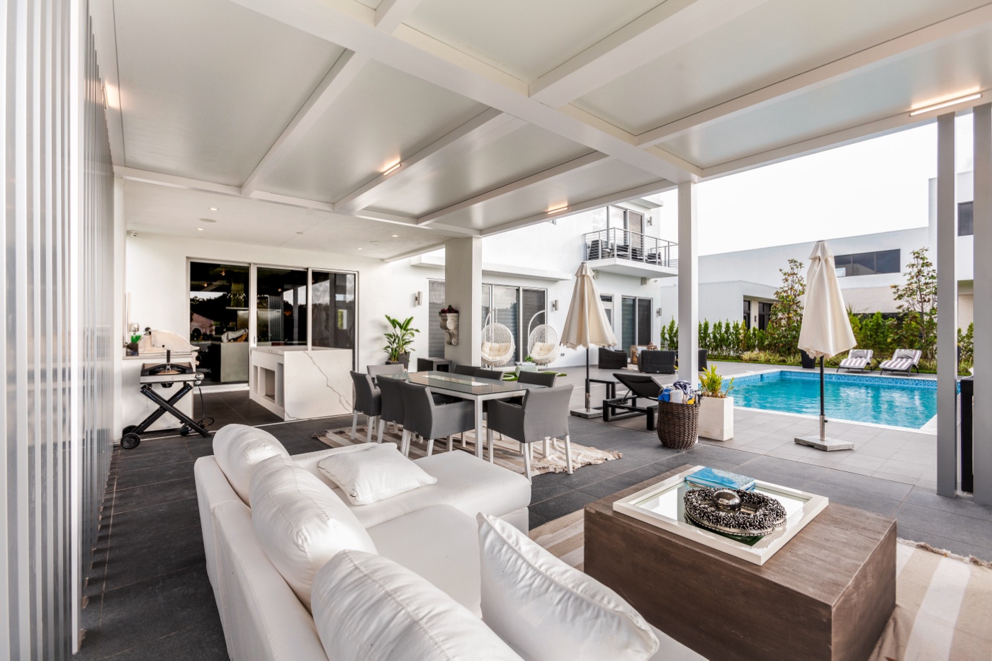 White pergola-covered outdoor kitchen and bar with grill, ceiling fan, and stone counter in a wooded yard