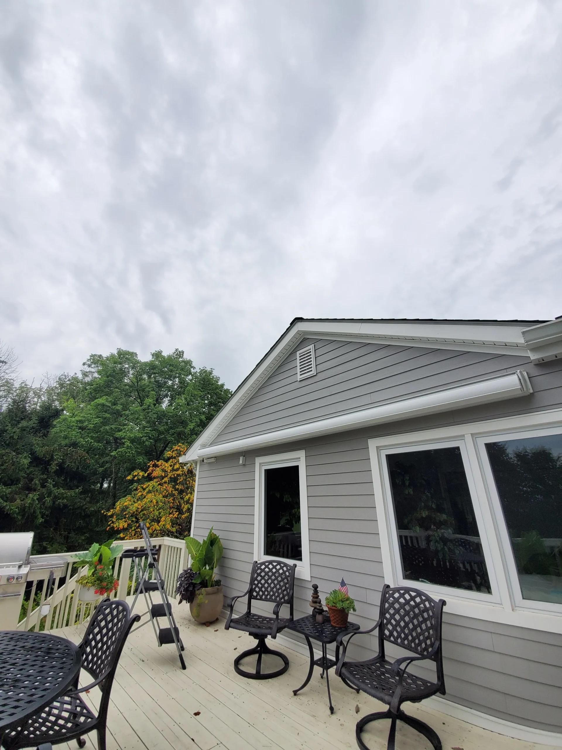 Closed retractable awning cassette mounted above windows on a gray home overlooking a deck with outdoor seating and surrounding greenery.