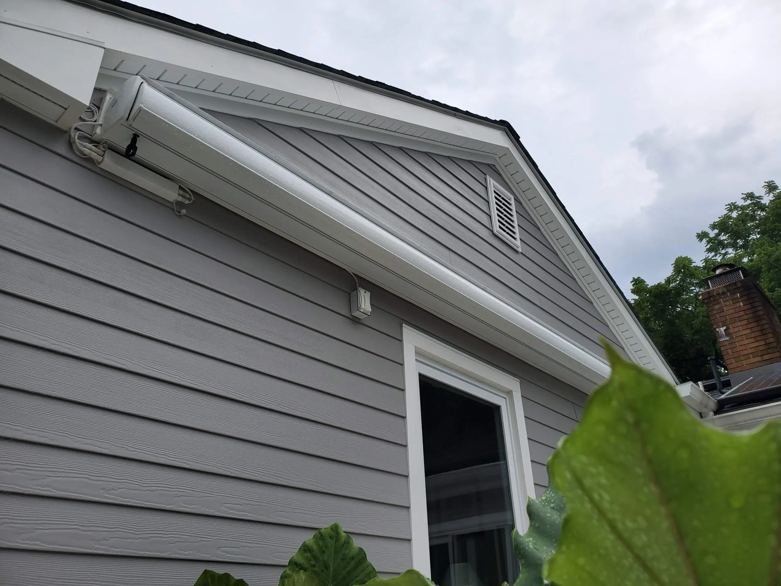 Closed retractable awning cassette mounted above a patio door on a gray home with surrounding greenery and a gabled roof.