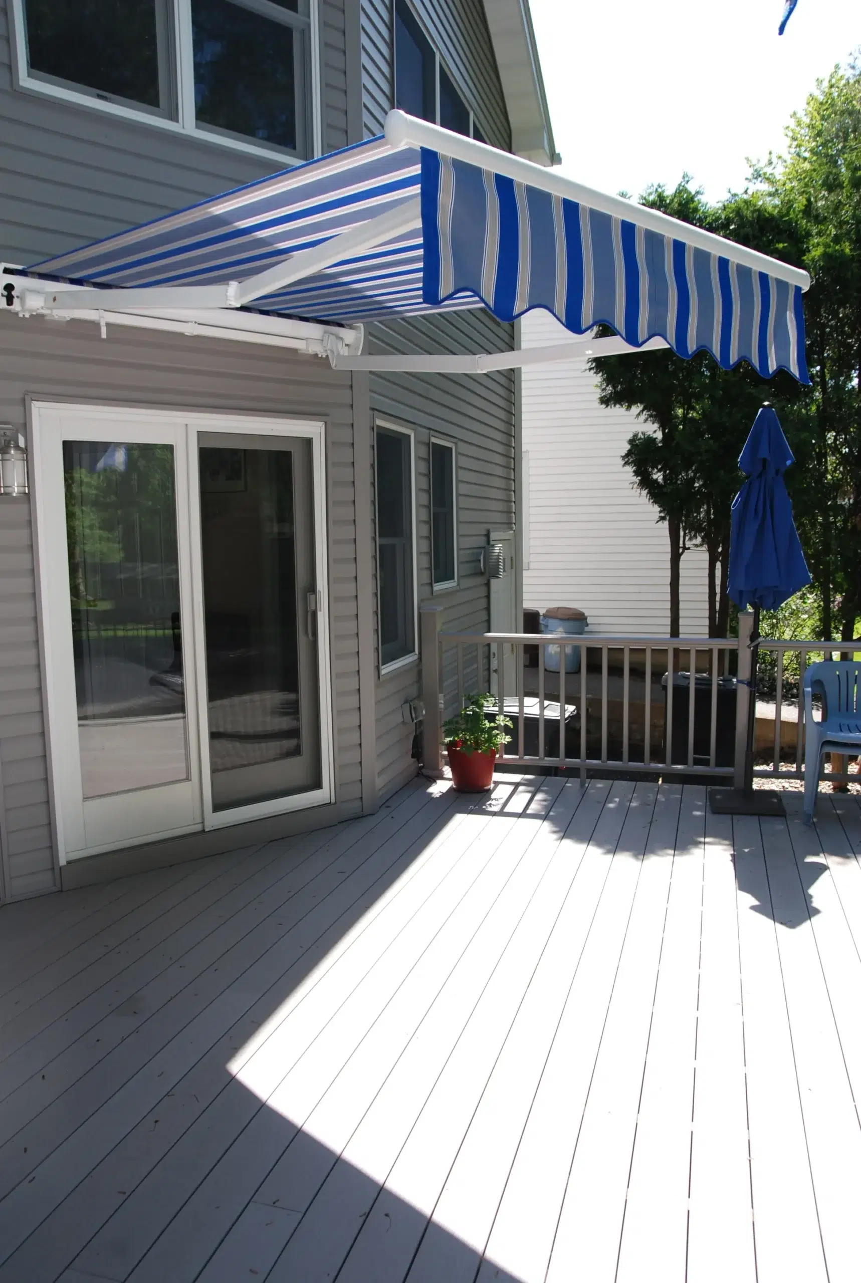 Blue-and-white striped retractable awning extended over a wooden deck outside a gray home with sliding glass doors.