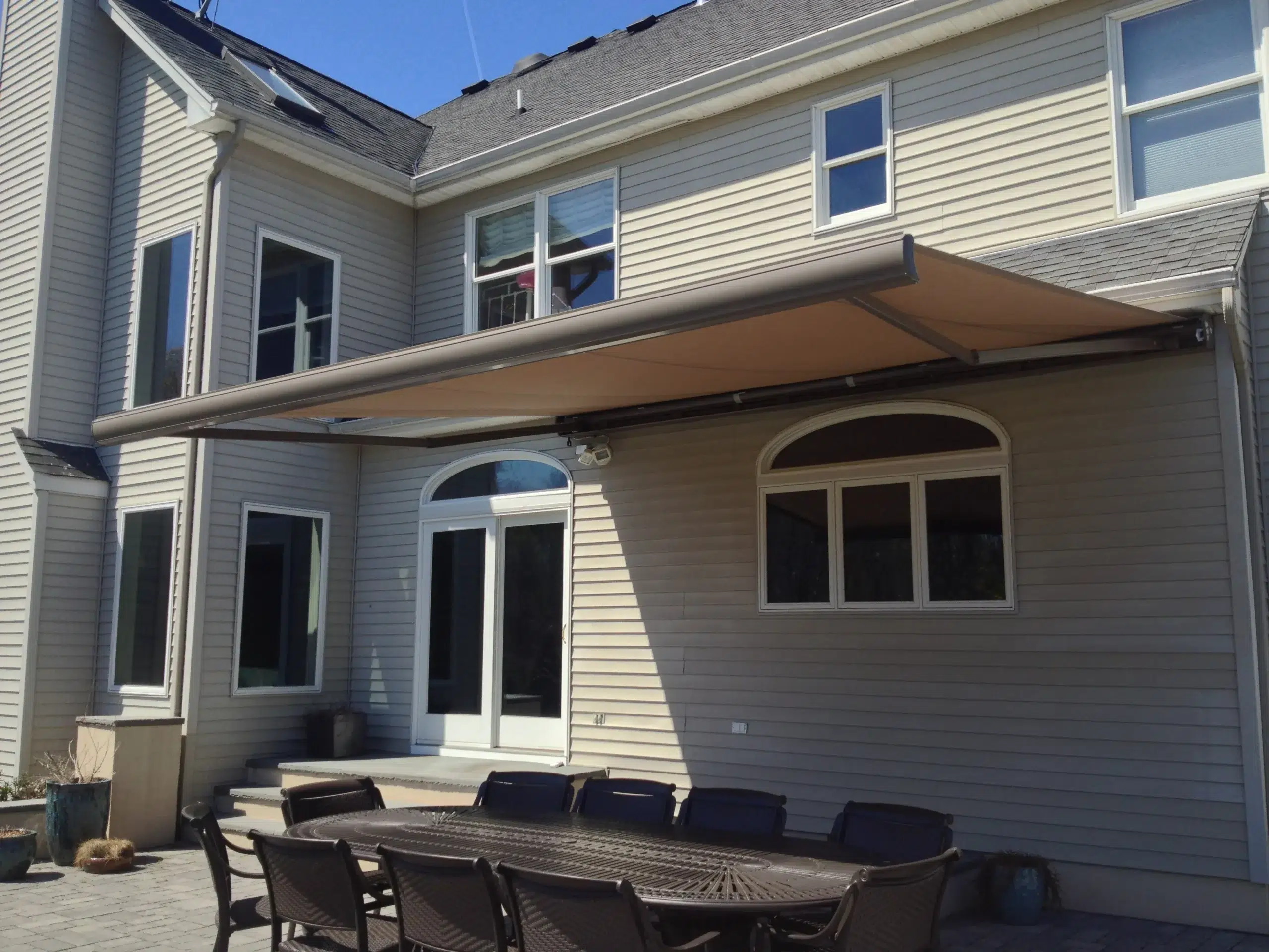 Retractable awning extended over a backyard patio with a long outdoor dining table attached to a beige two-story home.
