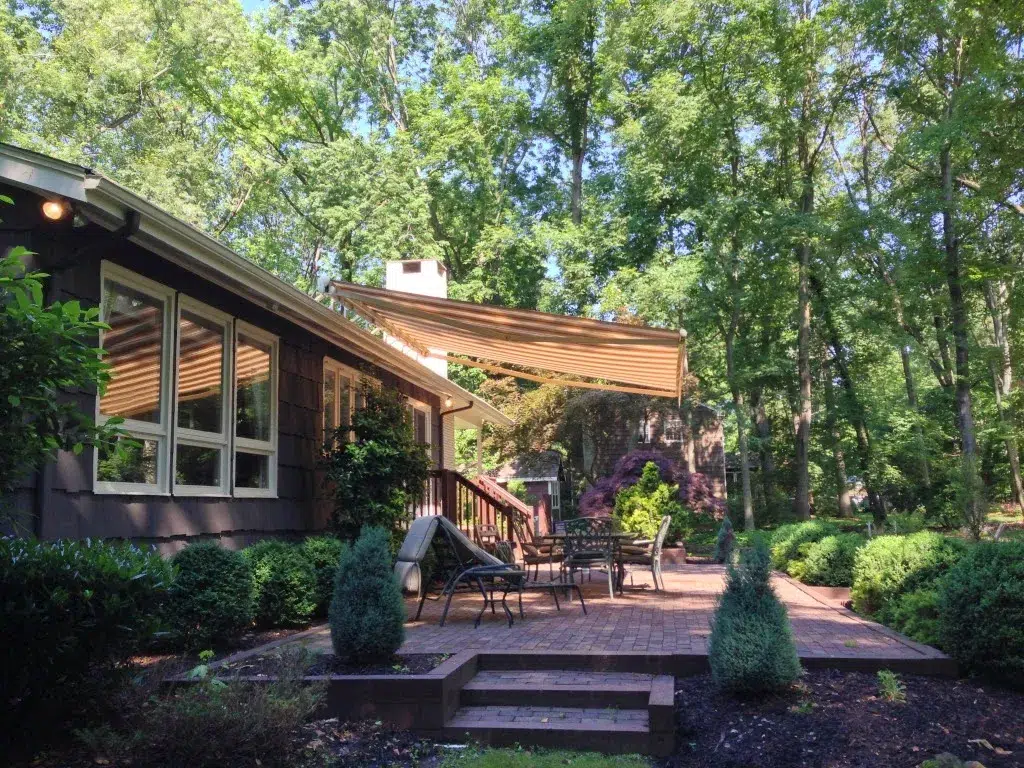 Retractable awning extended over a wooden backyard deck surrounded by lush greenery and outdoor seating.