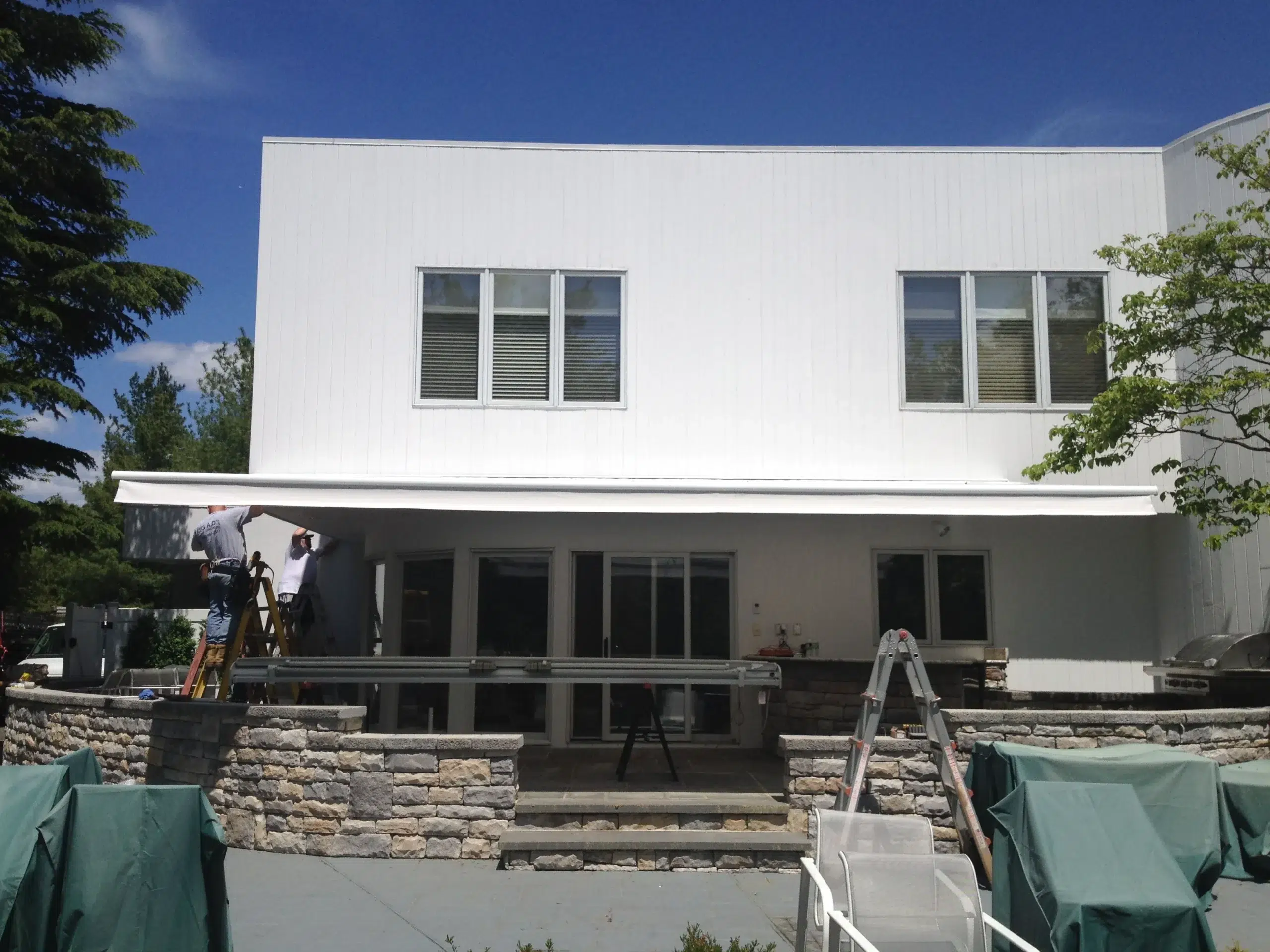 Retractable awning extended across the patio of a modern white two-story home with stone accents and outdoor seating.