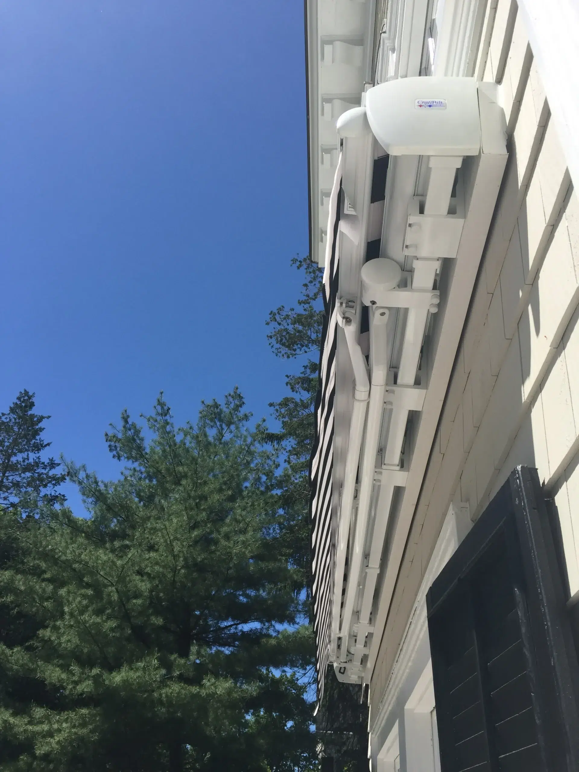 Close-up view of a retractable awning cassette and support frame installed on a home’s exterior wall with trees and blue sky in the background.