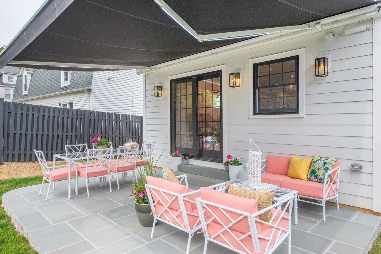 Dark retractable awning extended over a bright patio with pink cushioned seating, outdoor décor, and a white home exterior.