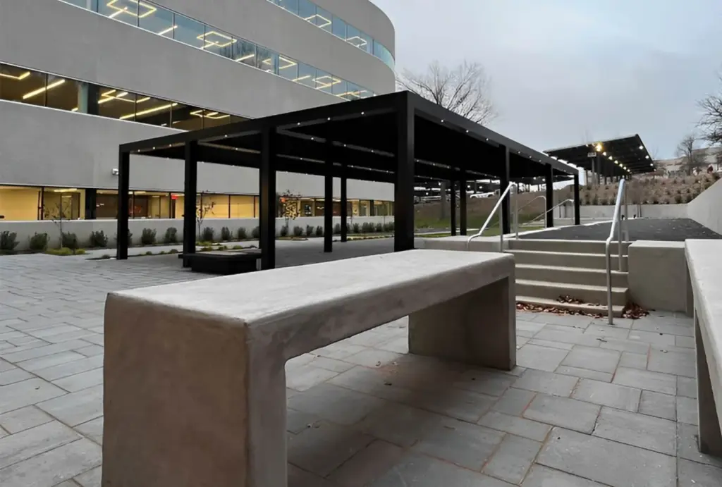 A modern outdoor space featuring a concrete bench, black canopy, and steps leading to a landscaped area beside a contemporary building.