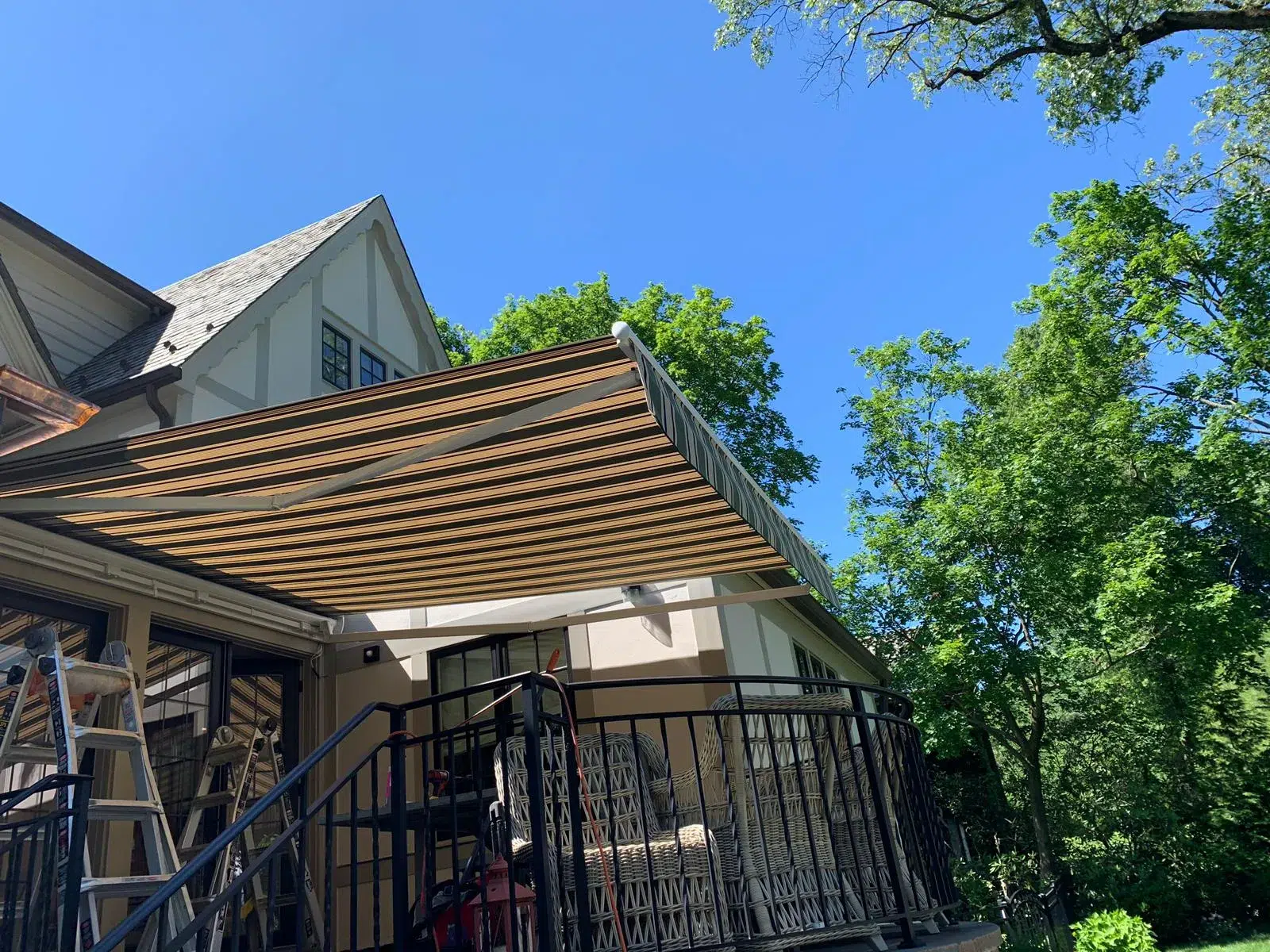 Retractable awning being installed above a raised backyard deck with black railings, surrounded by trees and greenery.