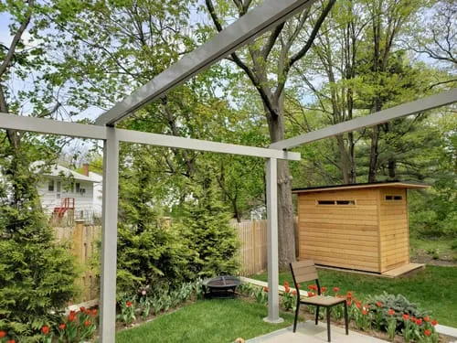 Modern pergola frame structure installed in a green backyard with trees, flowers, and a wooden shed in the background.
