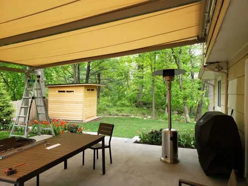 Retractable patio awning providing shade over an outdoor seating area with a table, heater, and garden view, including a small wooden shed in the background.