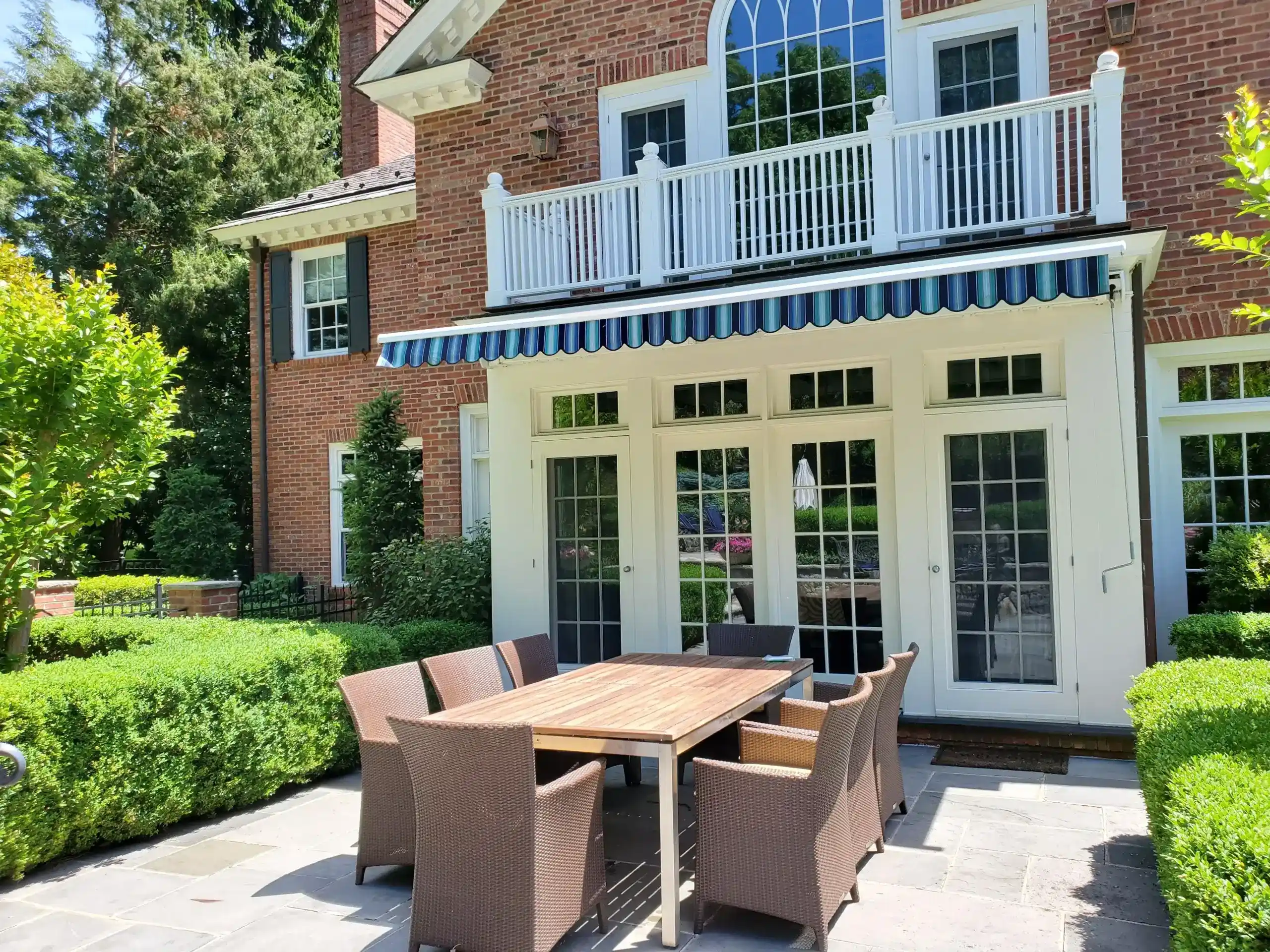 Blue-striped retractable awning shading the patio doors of a brick home with outdoor dining furniture in the garden.