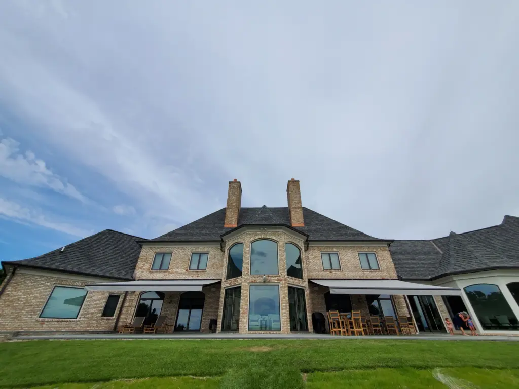 A grand two-story luxury home featuring retractable awnings over spacious patio seating areas, surrounded by lush green lawn and modern architectural design.