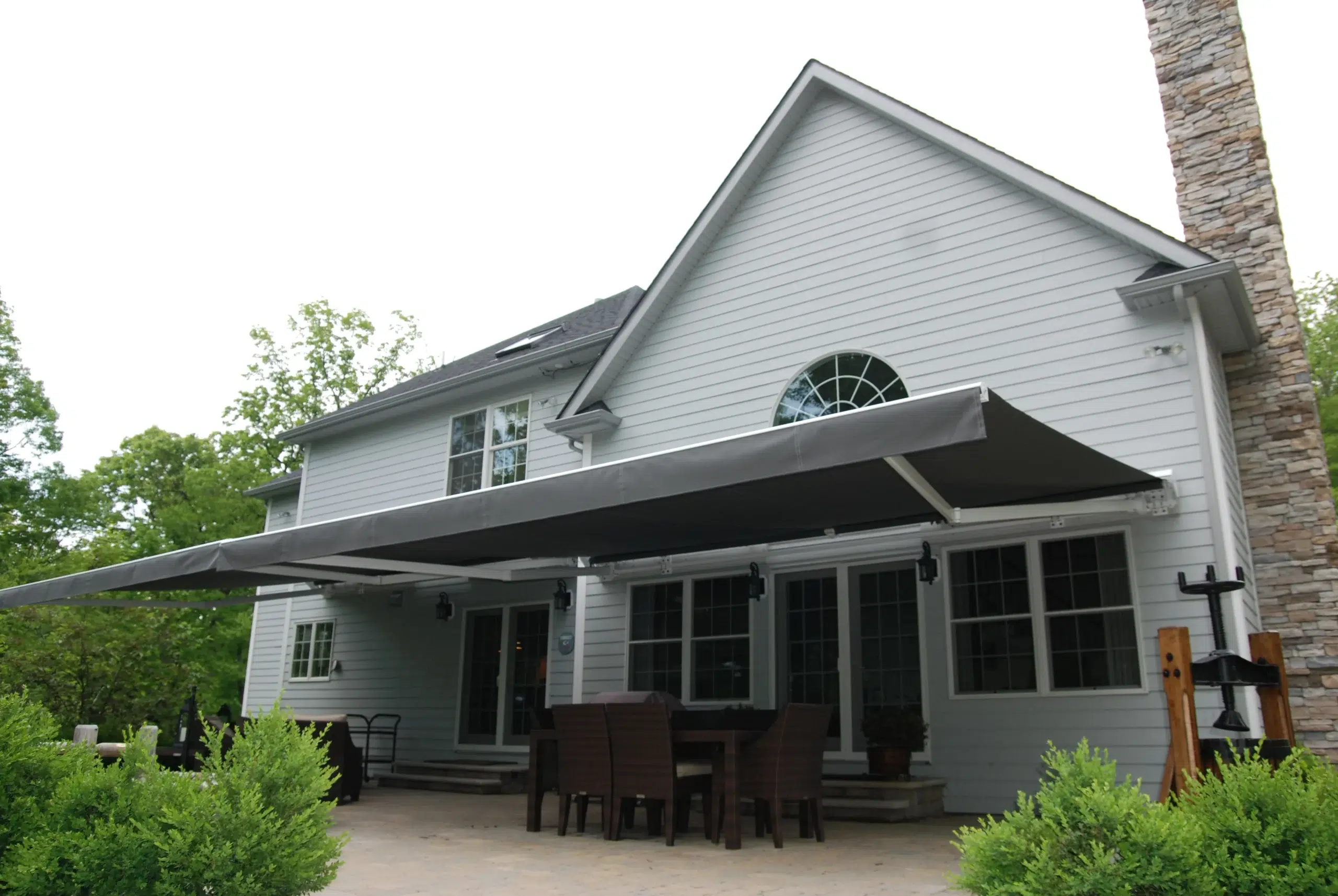 A large retractable awning covering an outdoor dining area beside a gray house, creating a shaded and elegant patio atmosphere.