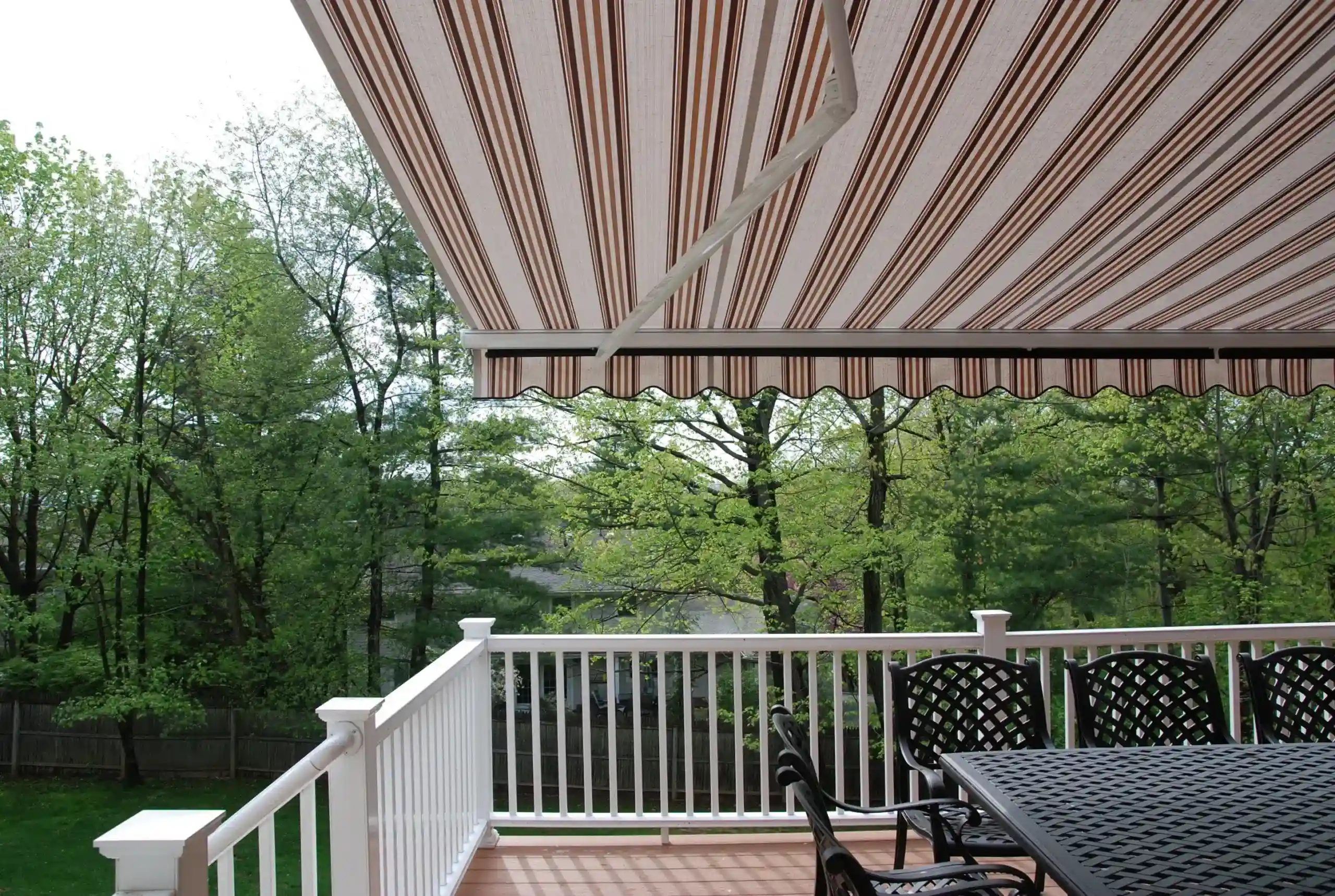 A retractable awning providing shade to a backyard patio dining area with black wrought-iron furniture and scenic greenery.