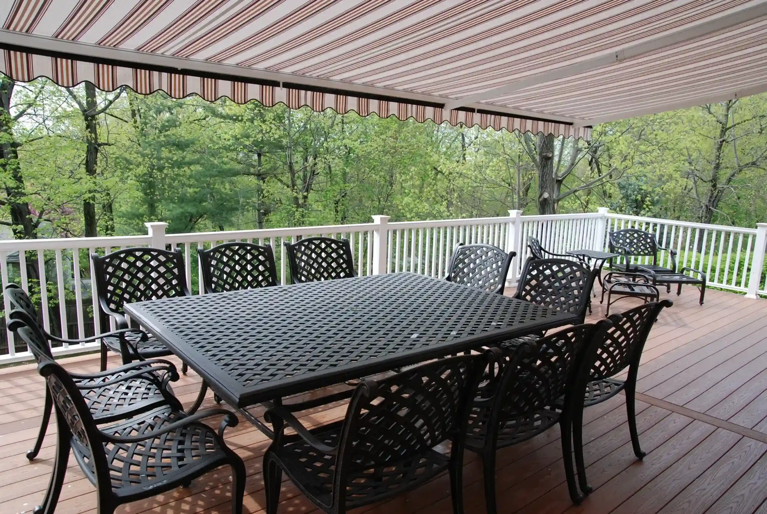 A striped retractable awning extending over a patio dining table and chairs on a wooden deck surrounded by trees.