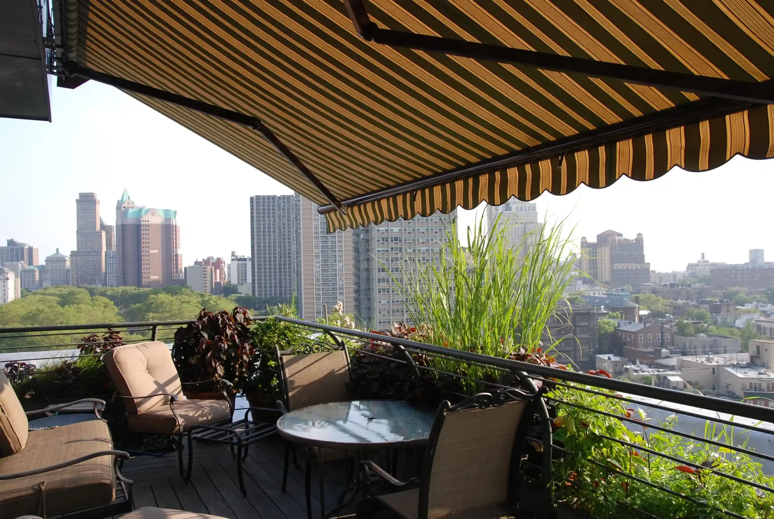 A yellow-striped retractable awning shading a furnished city balcony with outdoor seating and skyline views.