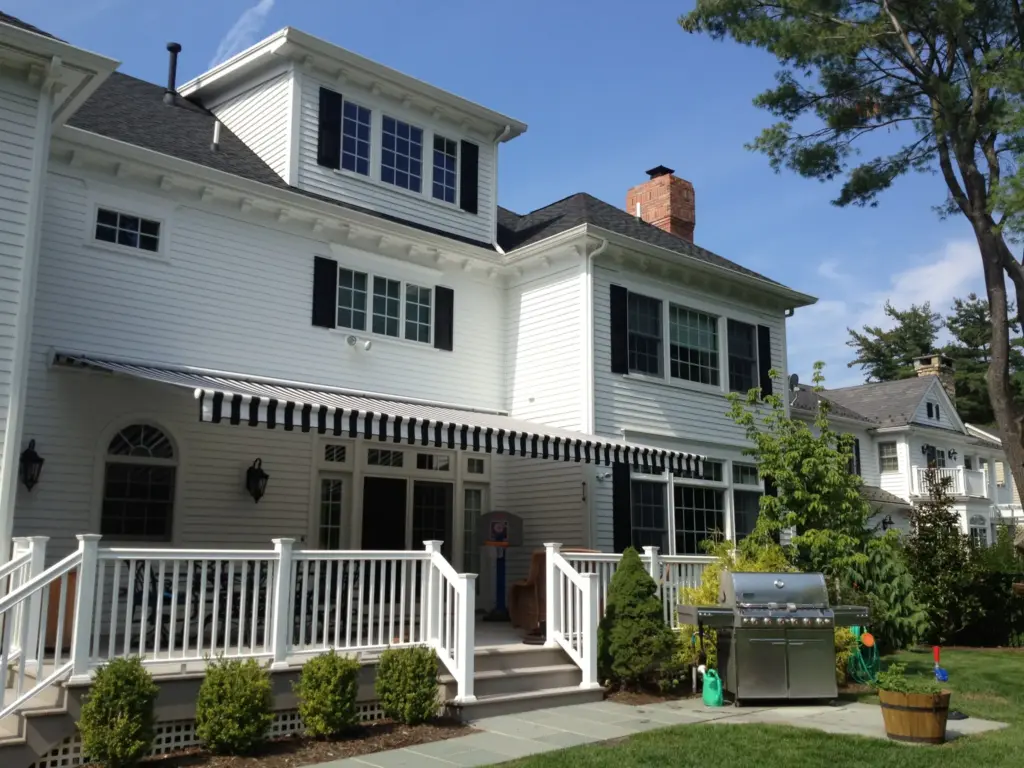 A striped retractable awning providing shade over a two-story white house patio with railing, outdoor furniture, and a well-kept garden.