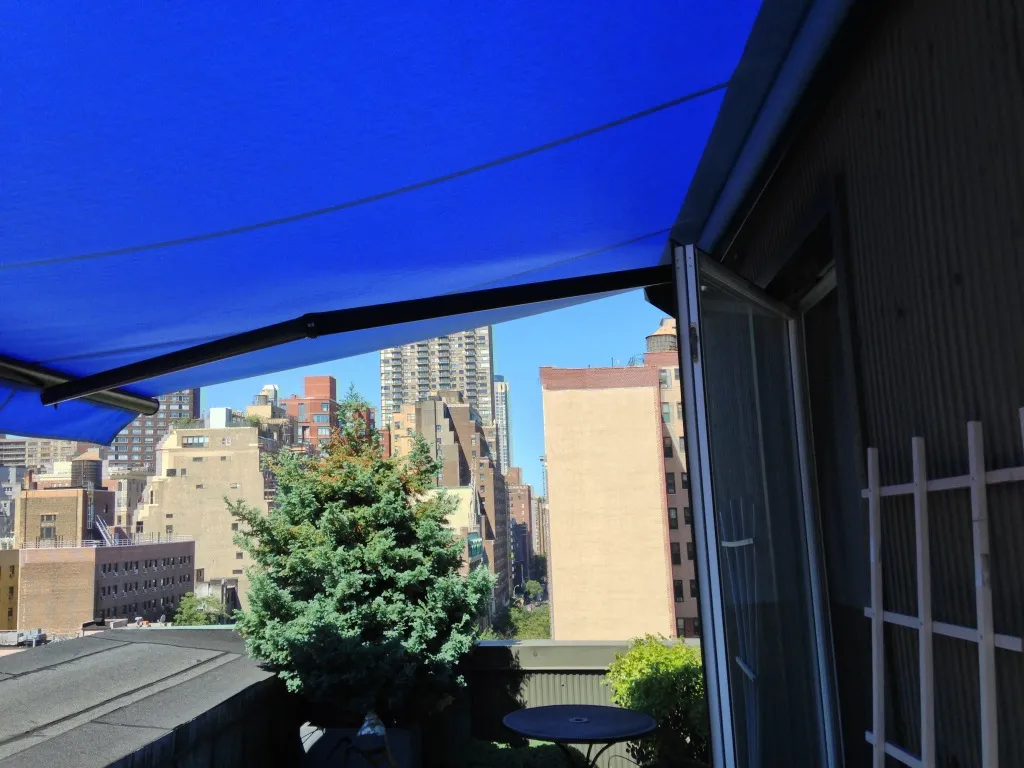 A bright blue retractable awning shading a rooftop patio with outdoor seating and a view of nearby city buildings.