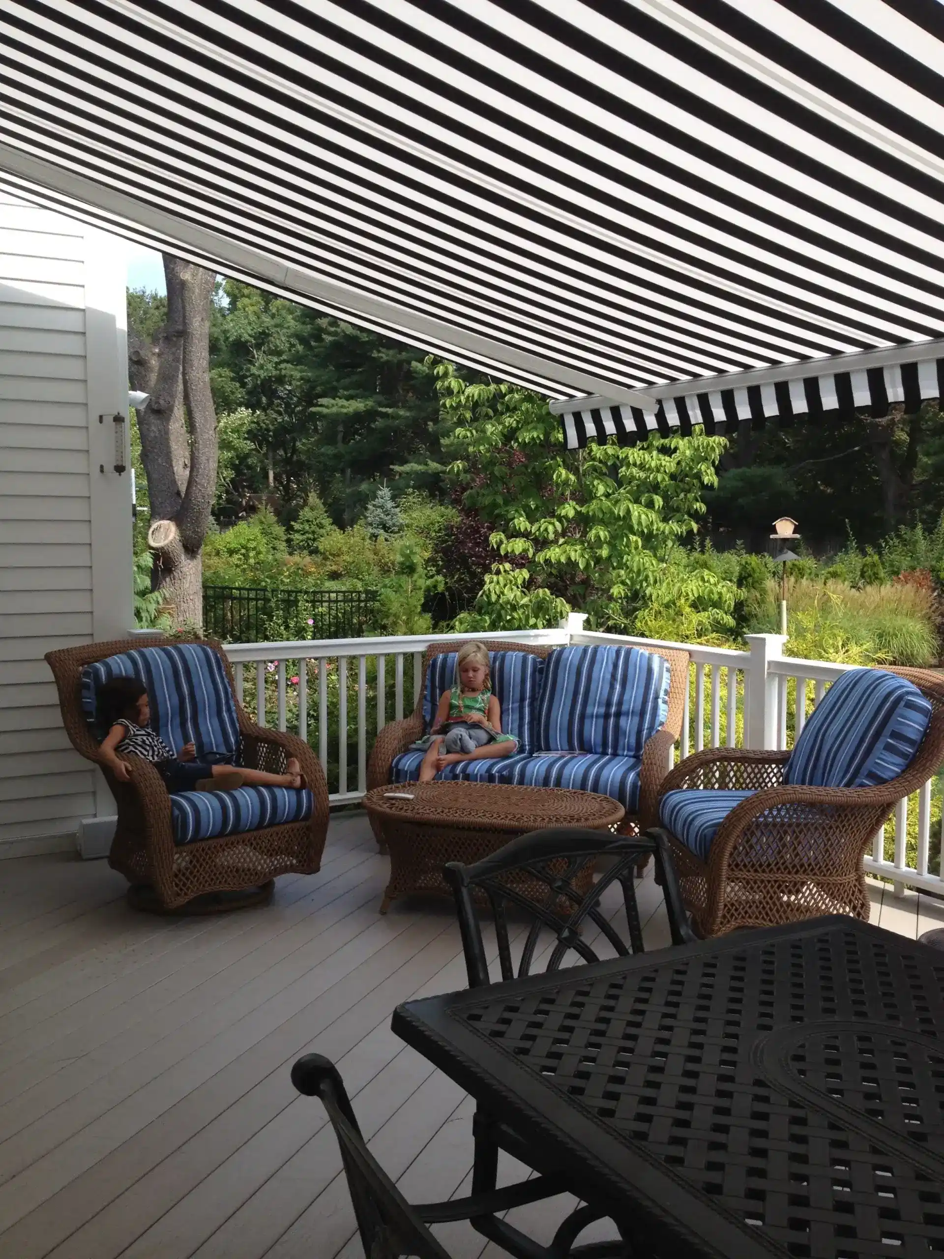 A shaded outdoor patio area with wicker furniture and blue cushions under a black-and-white striped retractable awning.