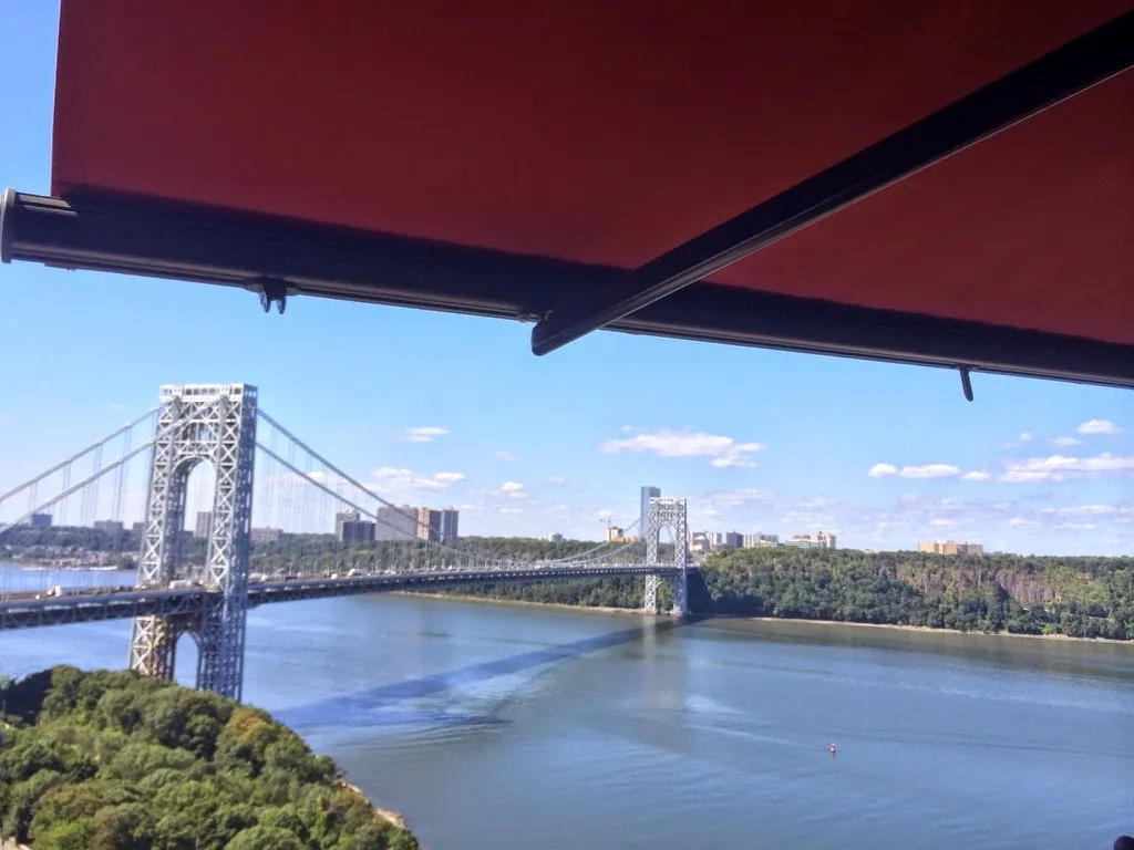 A red retractable awning providing shade to a balcony with a scenic view of a large suspension bridge and river landscape.