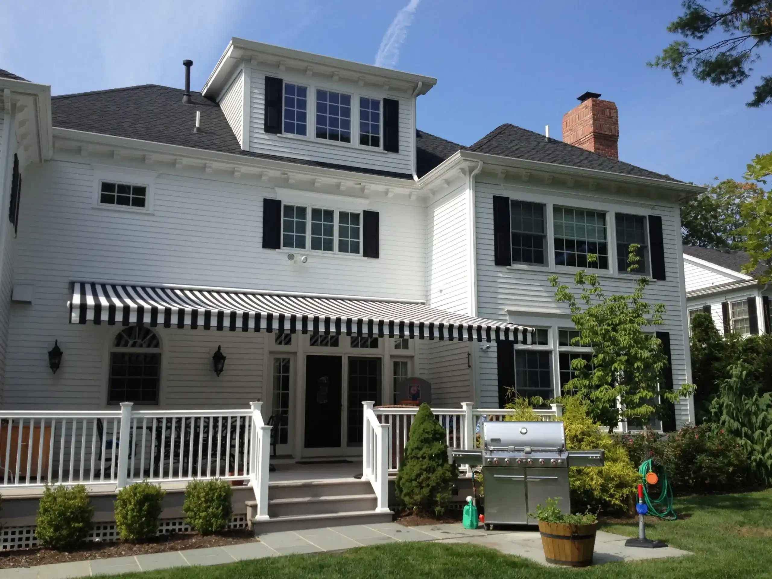 A black-and-white striped retractable awning extending over the patio of a white colonial-style home with railing and garden.