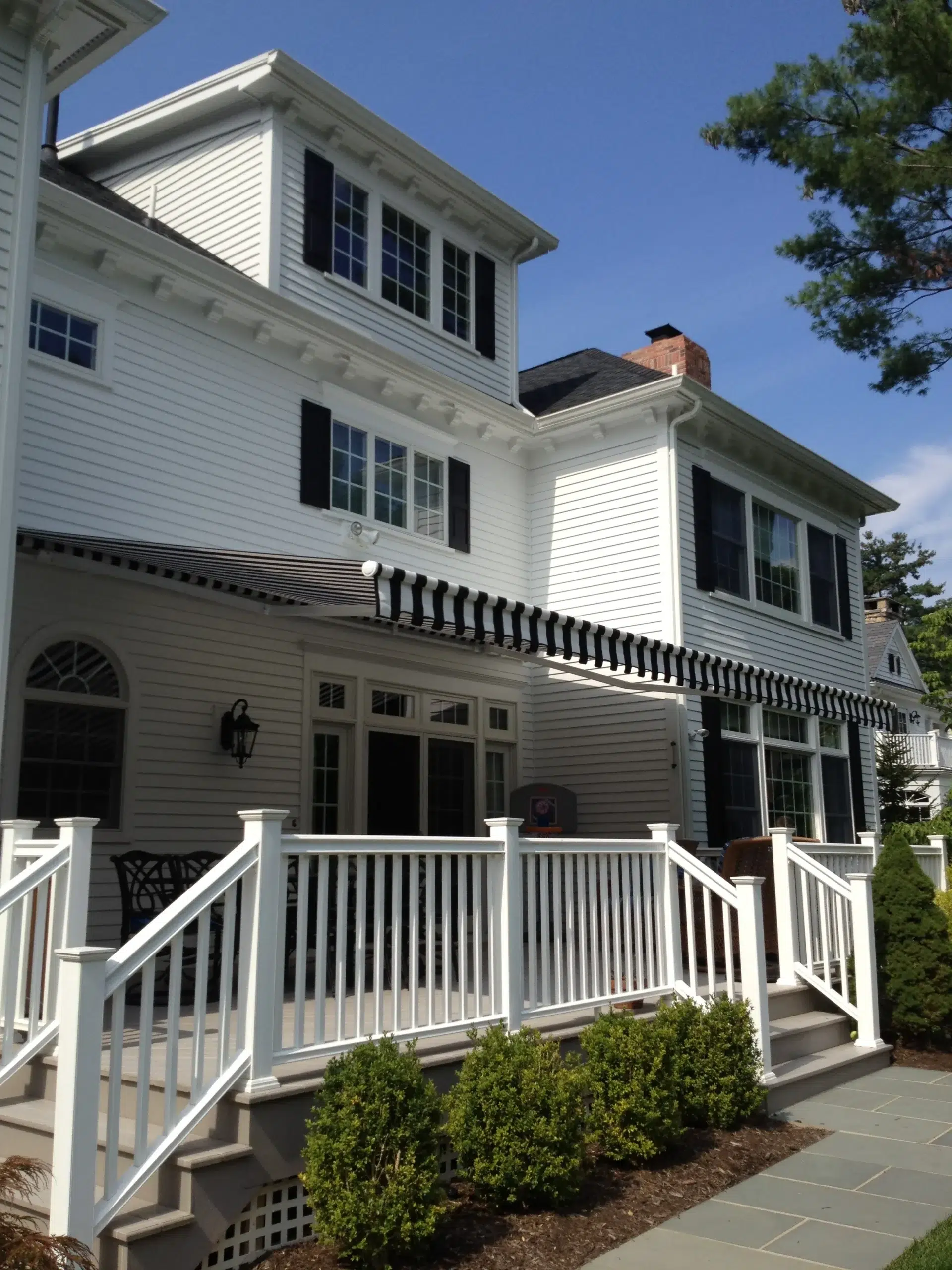A striped retractable awning mounted on a two-story white colonial home with a fenced patio and outdoor space beneath.