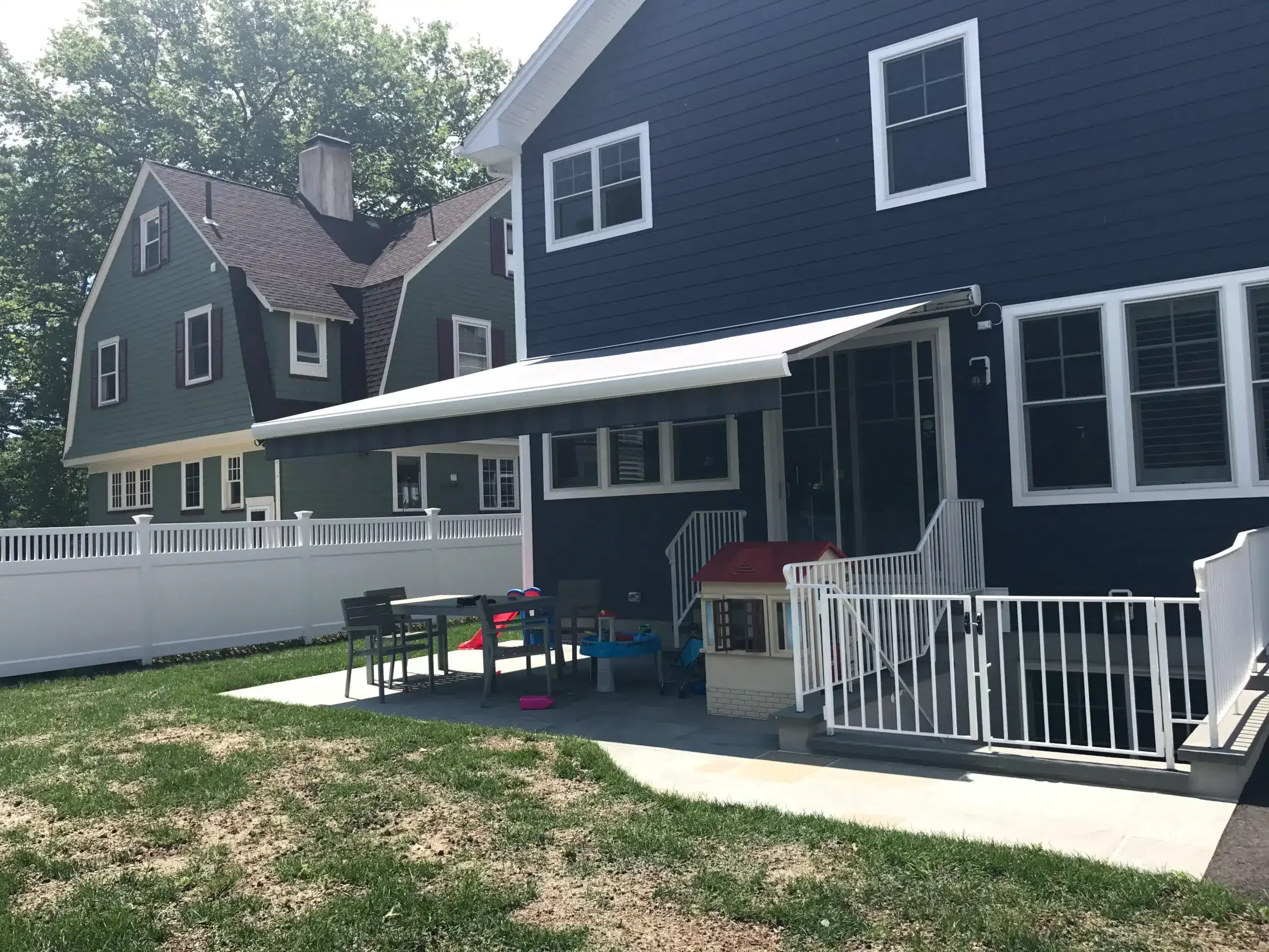 Retractable awning extended over a backyard patio of a dark-colored home with a fenced yard and outdoor seating.
