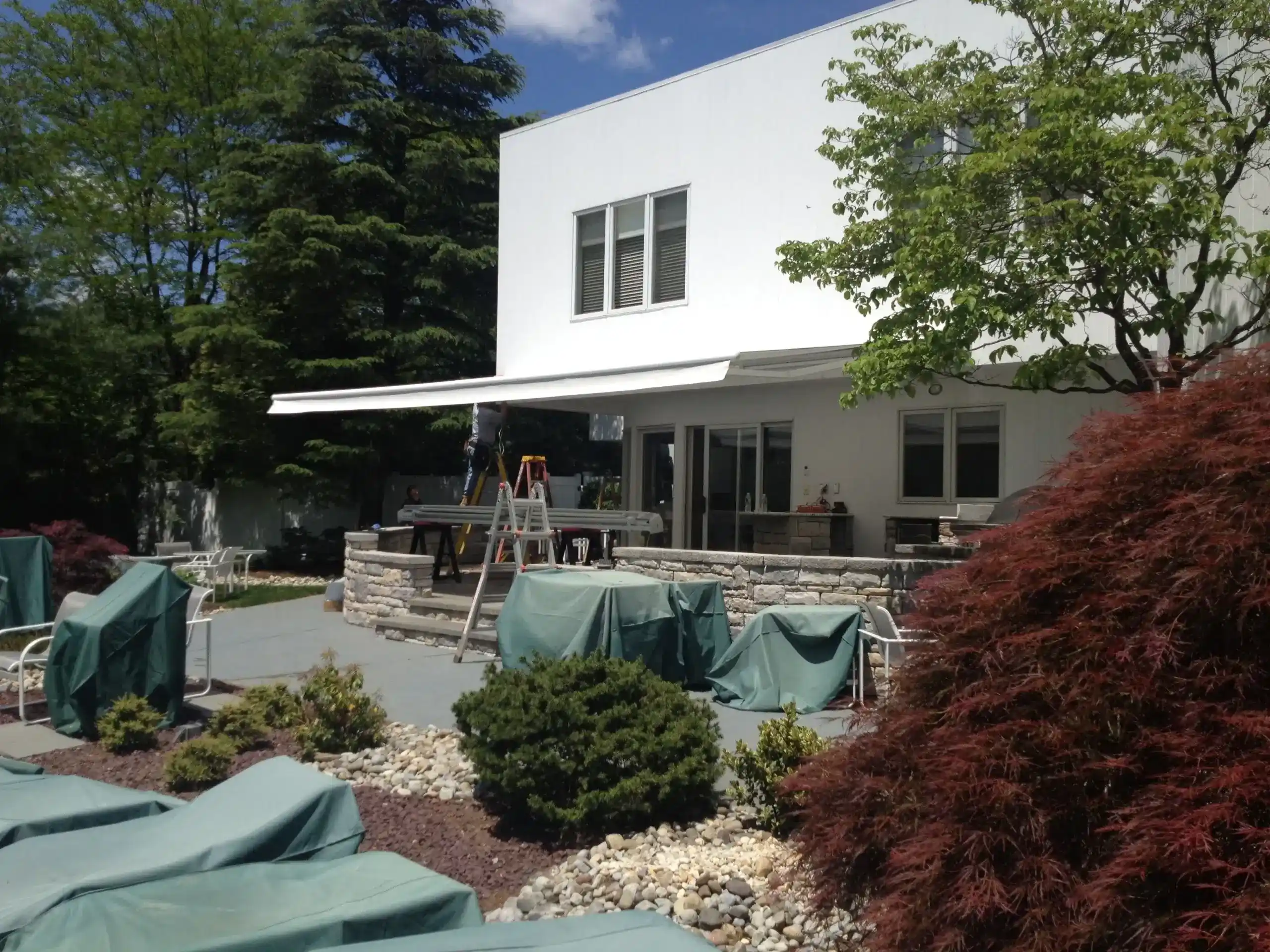 Retractable awning extended over the patio of a modern white home, surrounded by landscaped gardens and outdoor furniture.