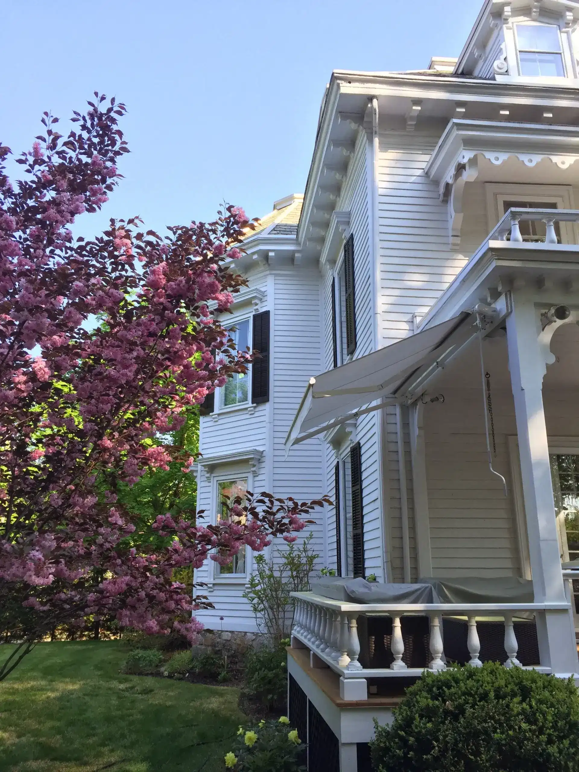Retractable awning extended over a traditional white porch on a historic-style home with blooming trees in the garden.