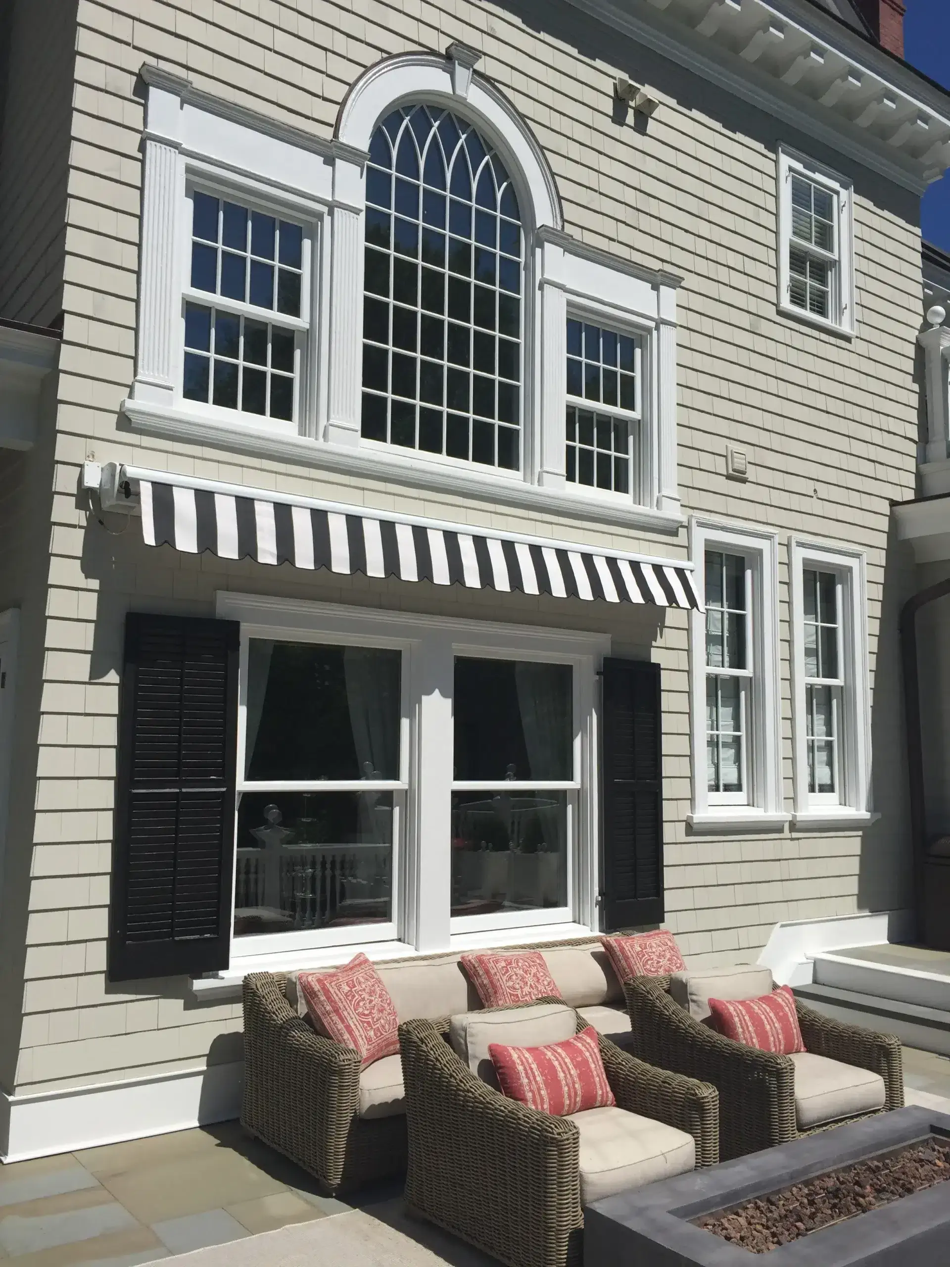 Black-and-white striped retractable awning installed above windows on a light-colored home with outdoor lounge seating below.