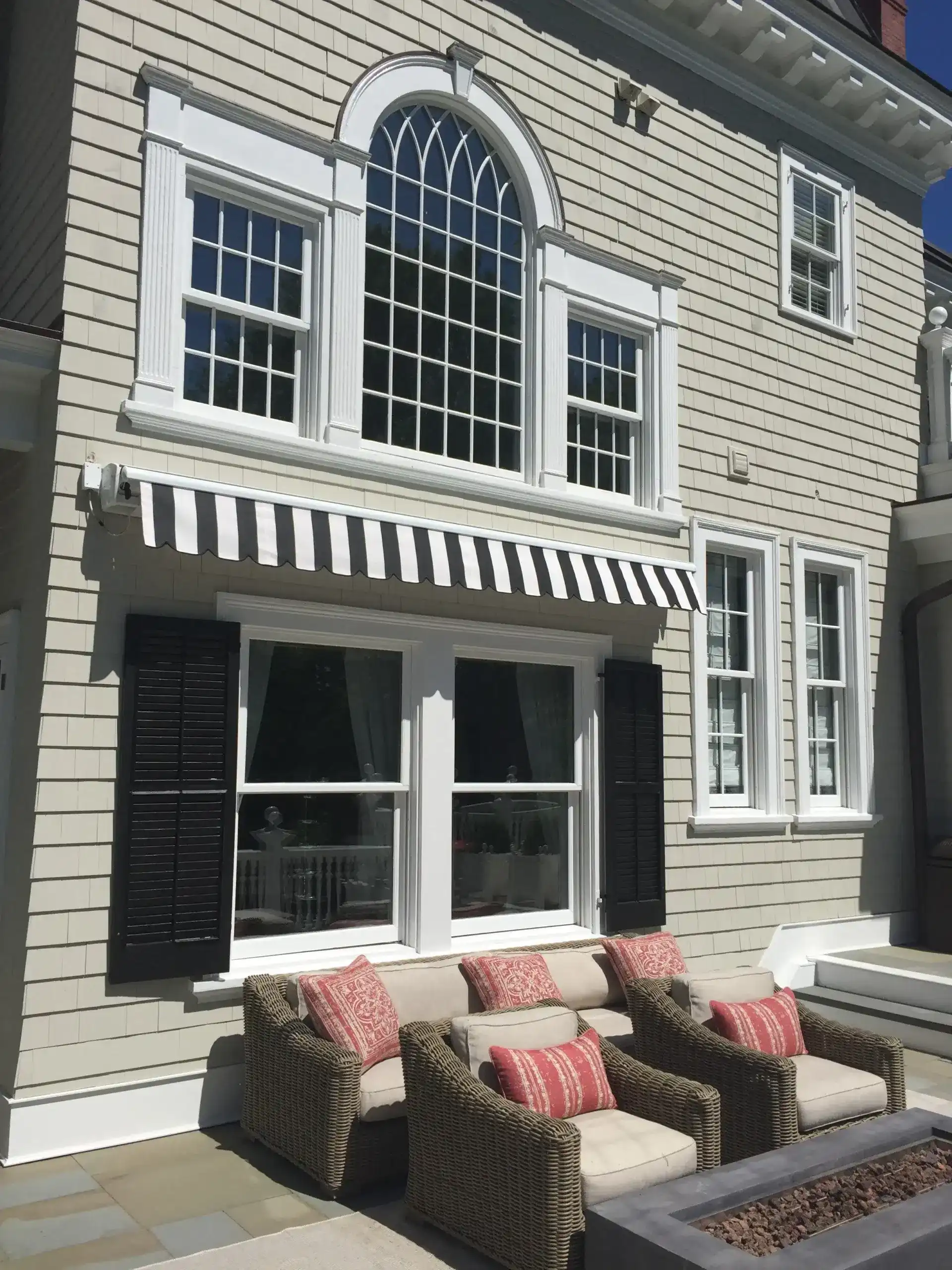 Black-and-white striped retractable awning installed above windows on a light-colored home with outdoor lounge seating below.