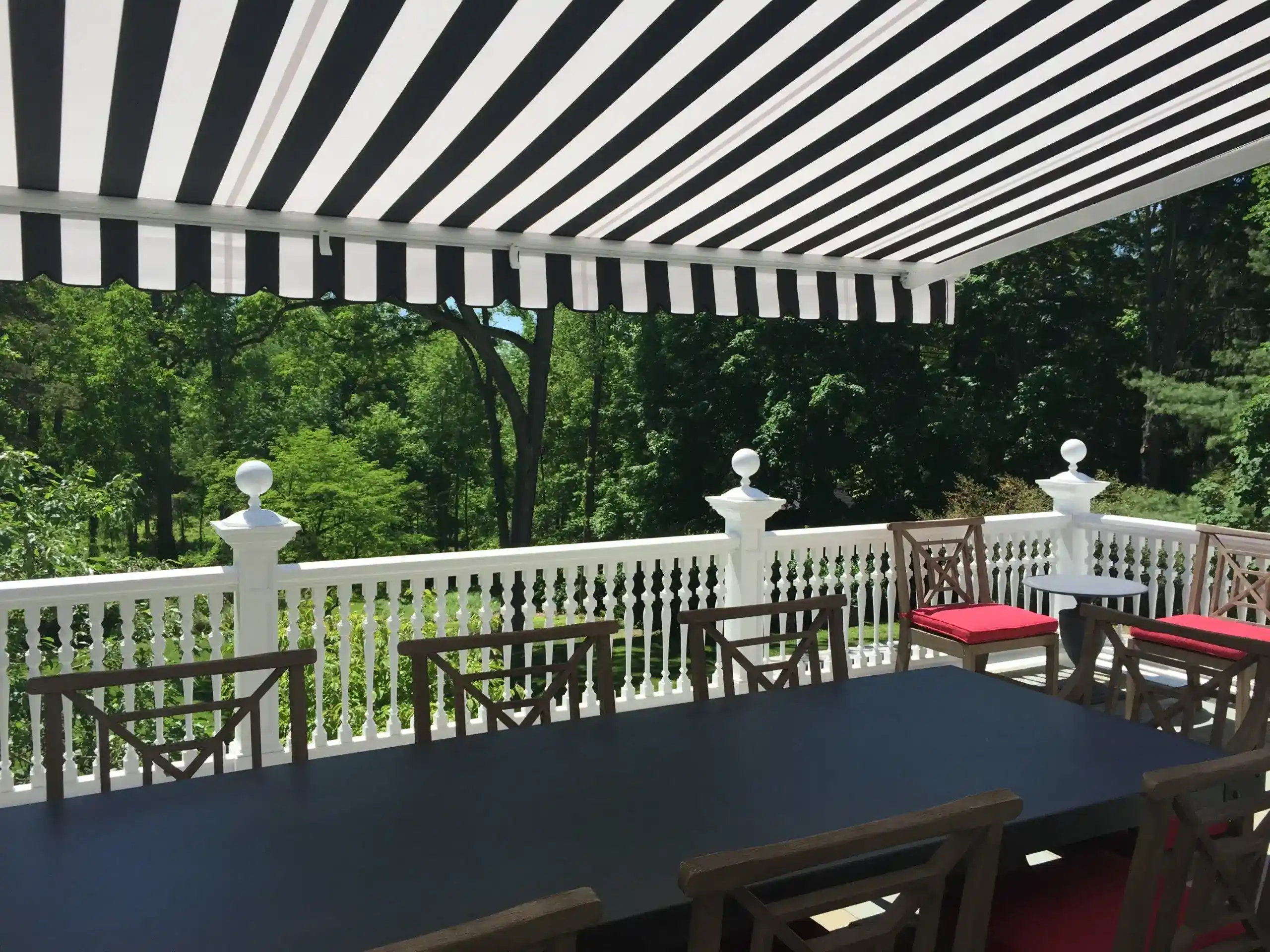 Black-and-white striped retractable awning providing shade over an outdoor dining table and chairs on a deck with scenic greenery.