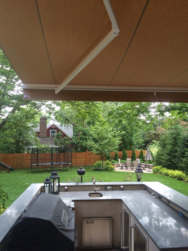 A retractable awning providing shade over a modern outdoor kitchen setup with a countertop and sink, overlooking a beautifully landscaped backyard with trees and a fence.