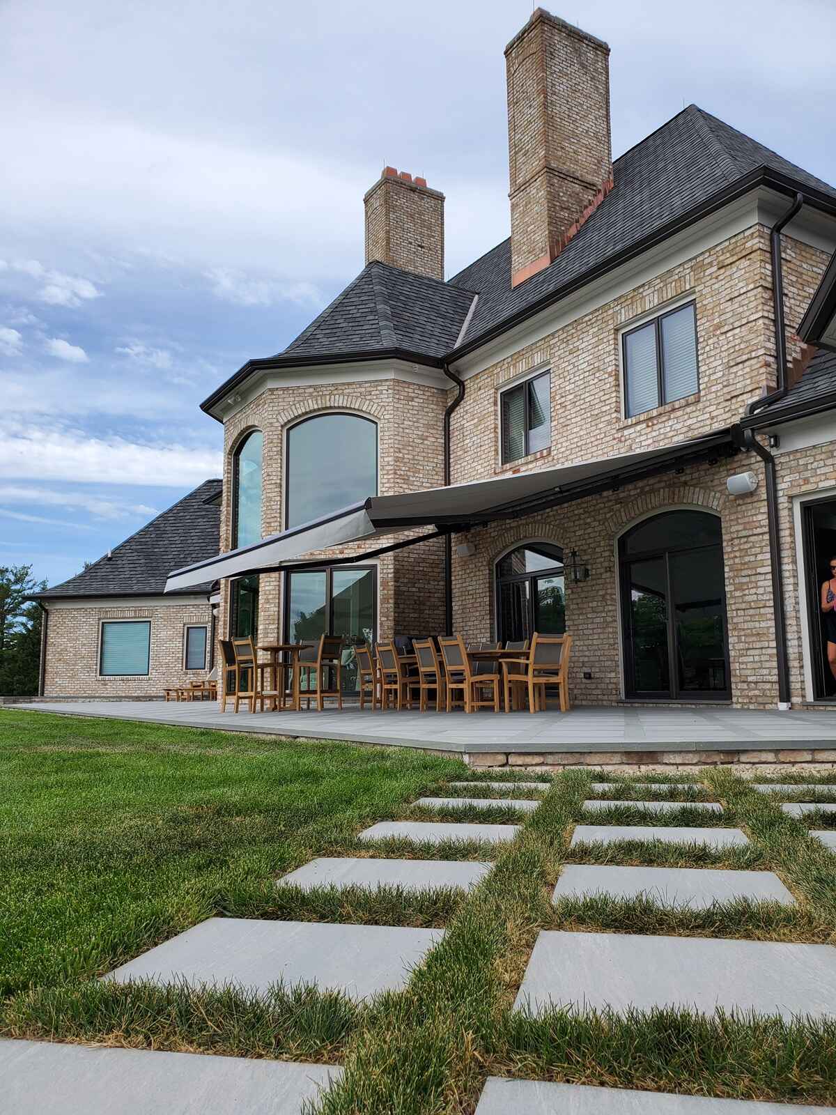 A modern retractable awning installed on a luxury brick home patio with a wooden dining set and stone walkway leading to a green lawn.