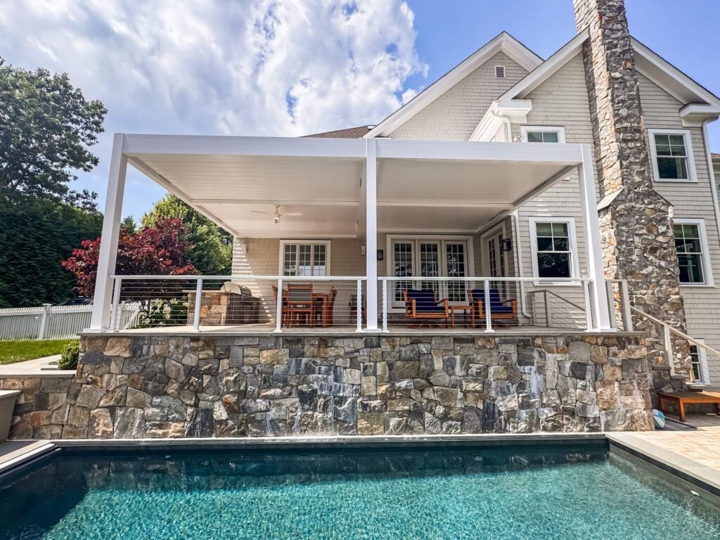 A modern poolside patio featuring a sleek white motorized pergola, stone wall design, and cozy outdoor seating, creating an inviting space under a bright blue sky.