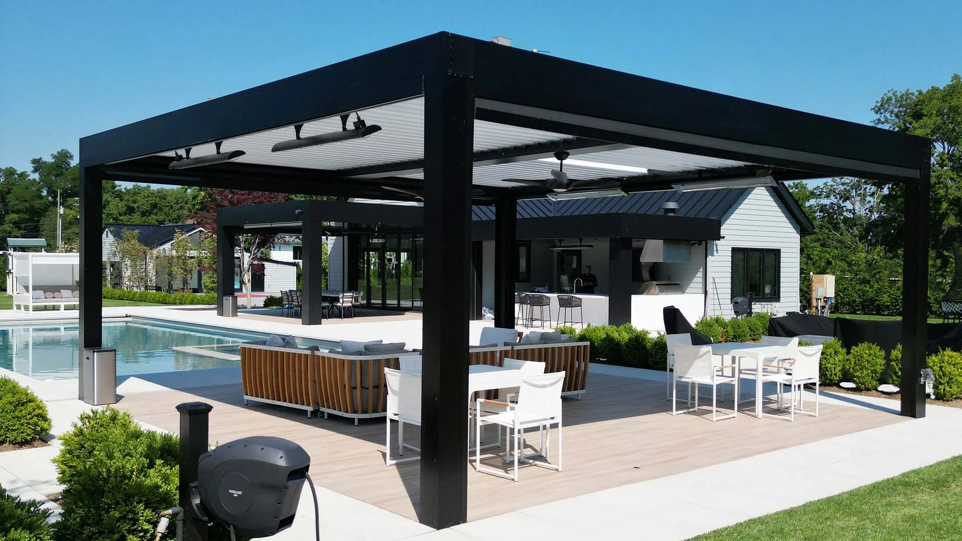 A sleek black louvered pergola installed beside a backyard pool, featuring outdoor dining and lounge seating in a modern residential setting.