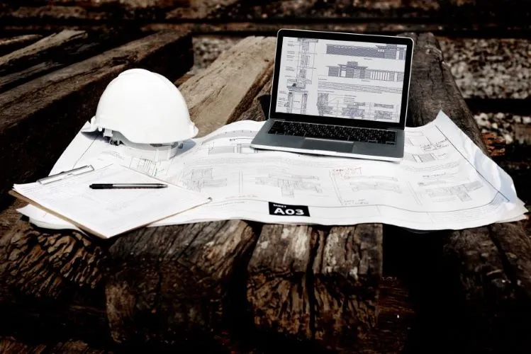 A laptop displaying construction plans placed on top of blueprints at a worksite, alongside a white safety helmet and clipboard.