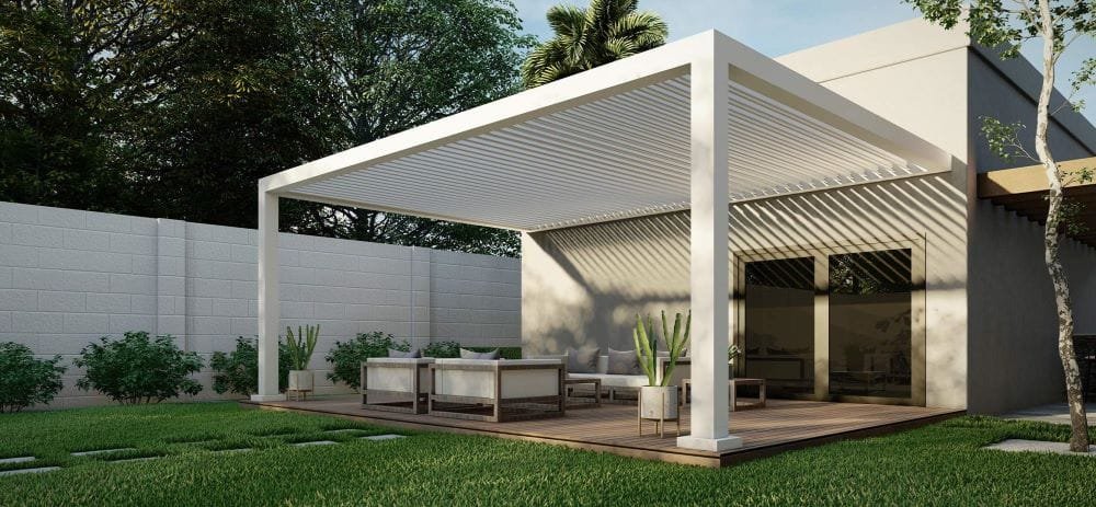 A sleek white louvered pergola installed over a backyard patio with outdoor seating, providing shade beside a modern home.